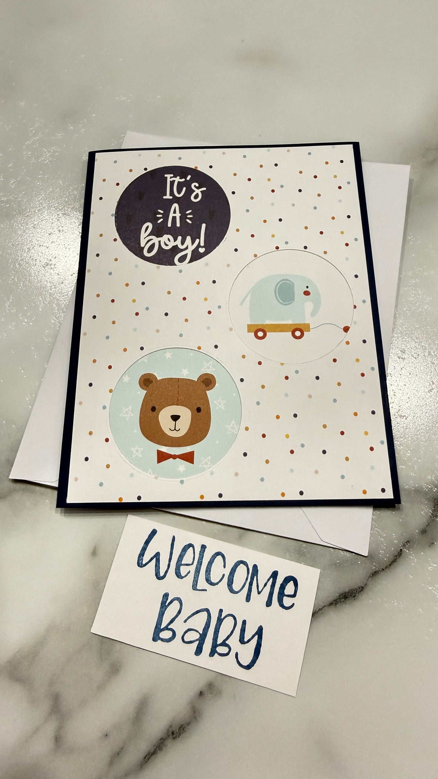 A handmade baby shower card with a 'It's a boy!' design featuring a teddy bear and camper illustration, displayed on a marble surface next to a small 'Welcome Baby' card.