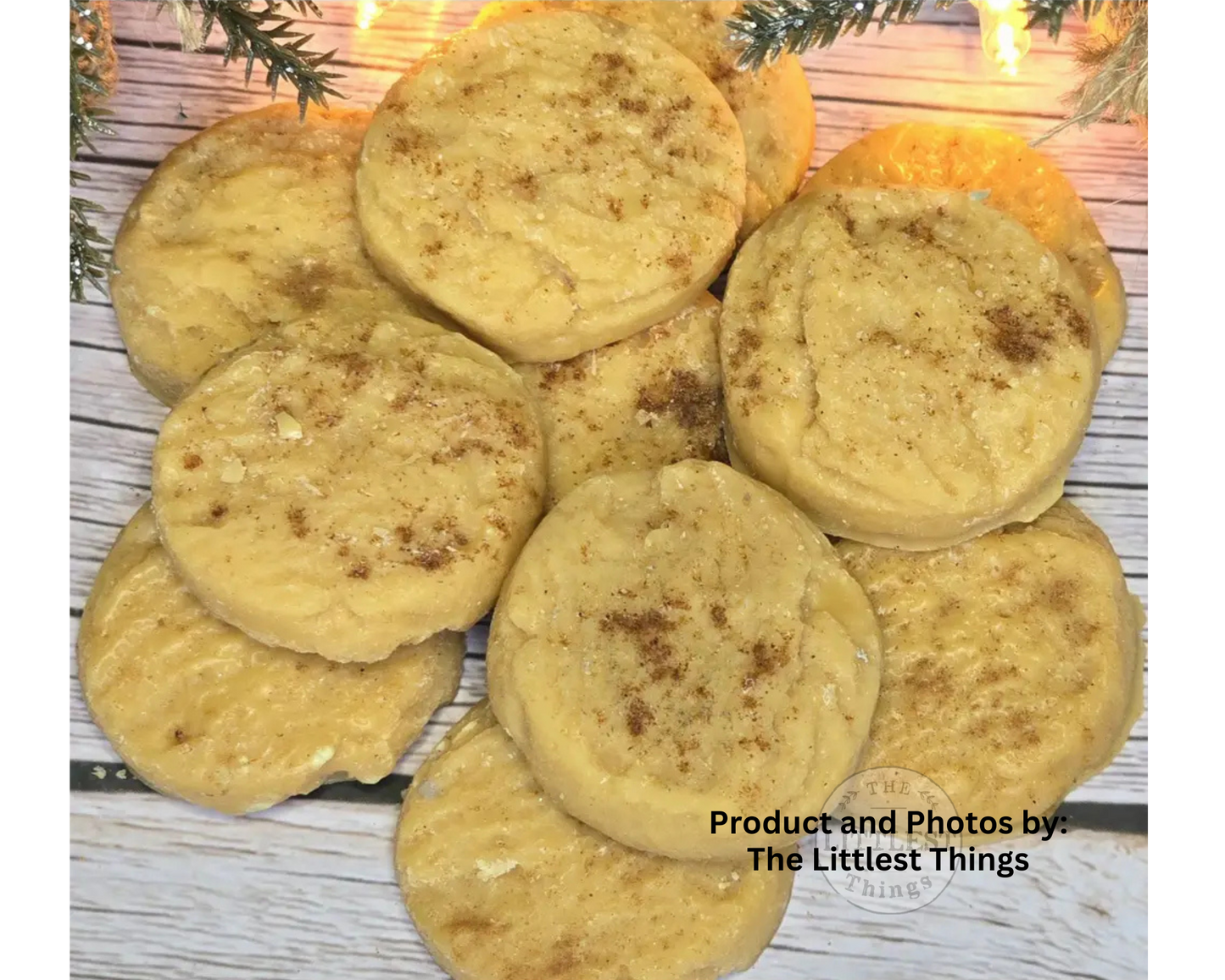 A pile of golden-brown sugar cookies sprinkled with cinnamon, arranged on a white-washed wooden surface with holiday greenery and warm string lights in the background.