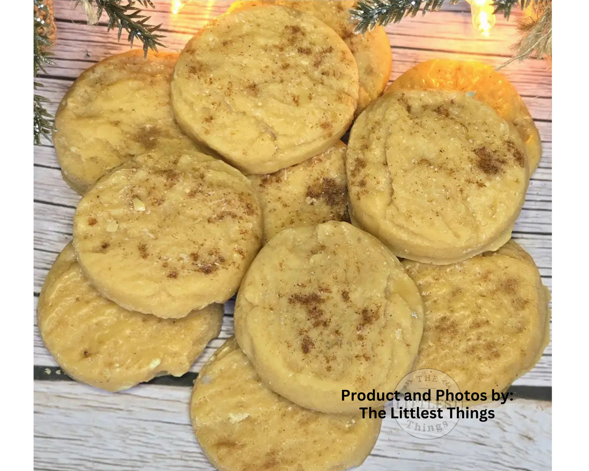 A pile of golden-brown sugar cookies sprinkled with cinnamon, arranged on a white-washed wooden surface with holiday greenery and warm string lights in the background.