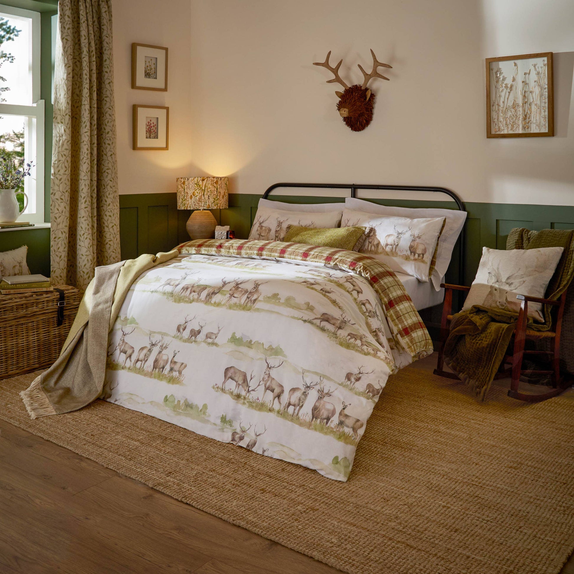 A rustic bedroom scene featuring a bed dressed in a deer-patterned duvet cover and matching pillow shams, styled with a plaid throw blanket. The room has a green and cream wainscoted wall, a wooden floor, a woven rug, and a rocking chair.