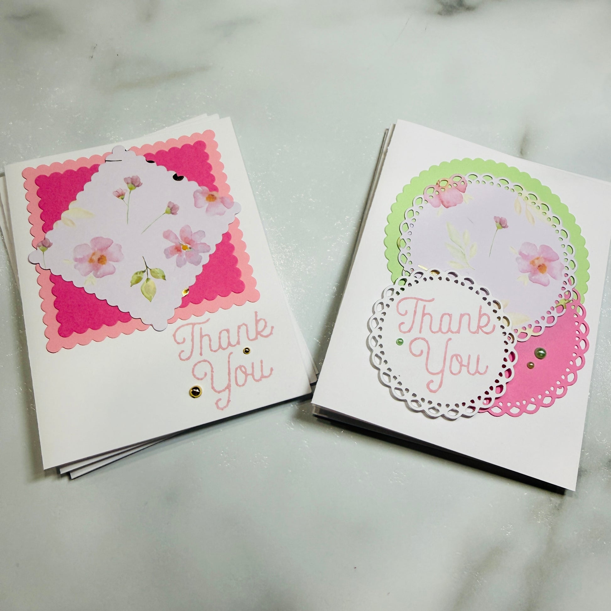 Two handmade thank-you cards displayed on a light marble surface. One card features pink and white layered paper with small flowers and the words 'Thank You' in pink script, while the other has white, pink, and light green layered paper with a lace doily and 'Thank You' text.