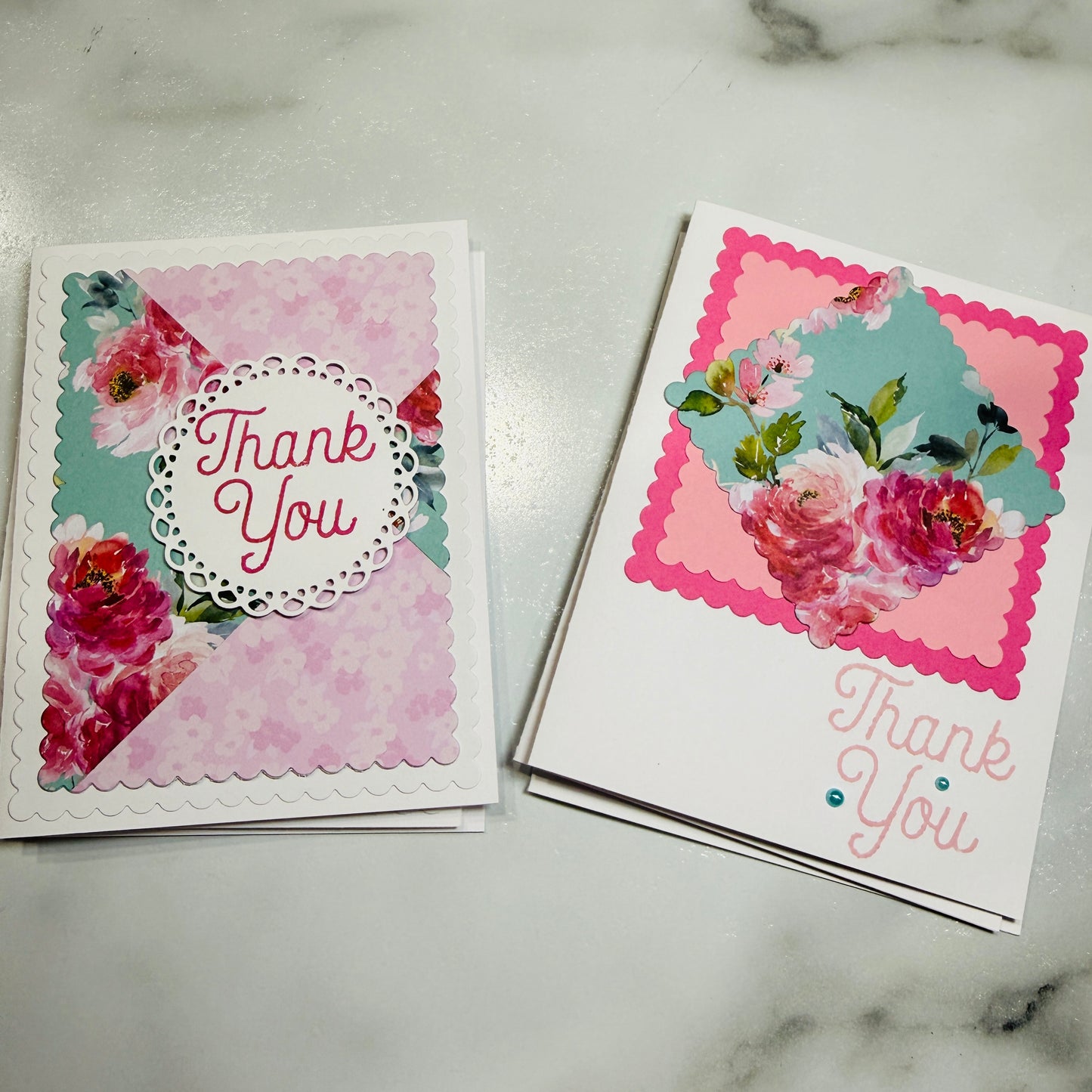 Two handmade floral thank-you cards displayed on a light marble surface. The card on the left features a pink floral pattern with a white scalloped circle containing the words 'Thank You' in pink.