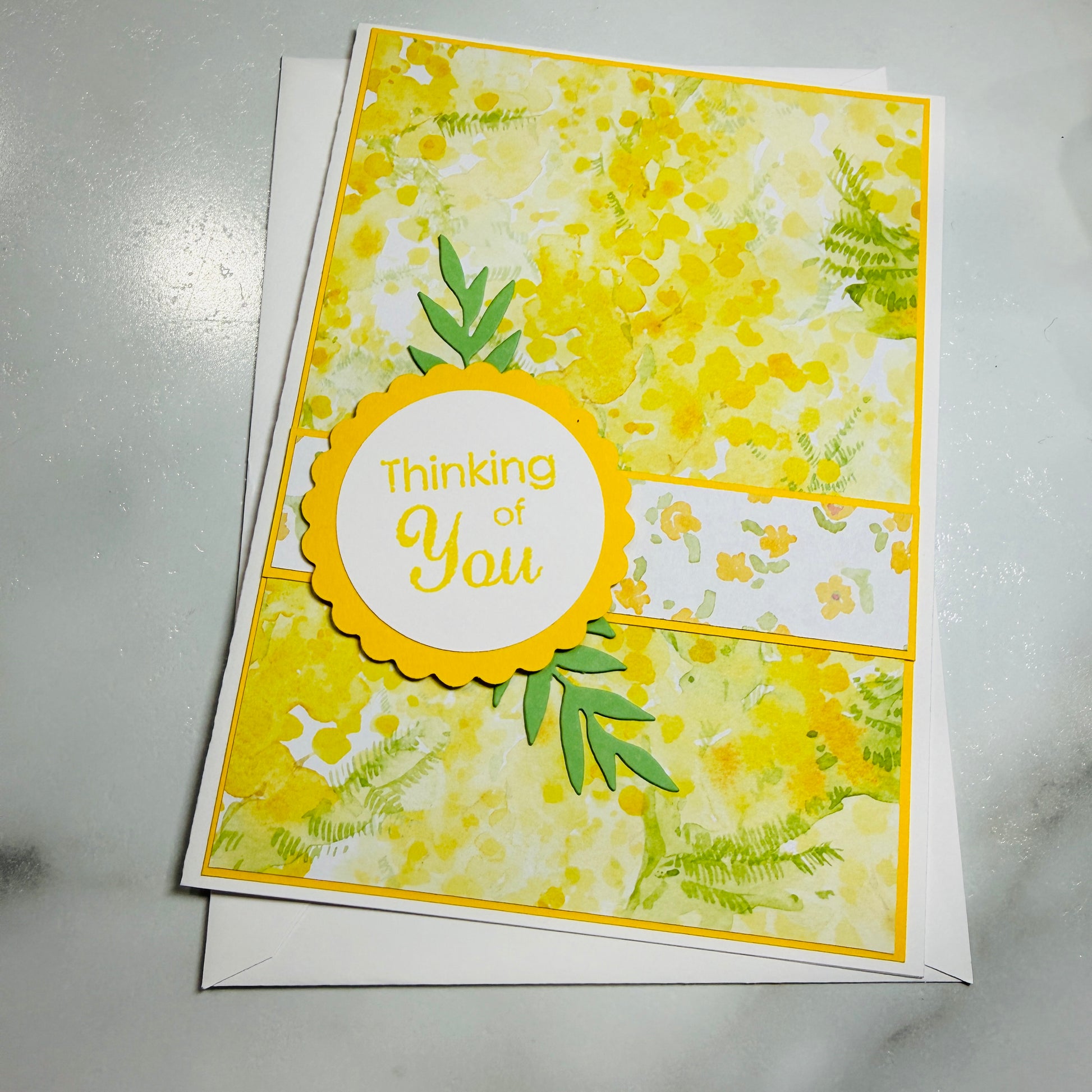 A handmade greeting card with a yellow and white floral pattern, featuring a scalloped yellow circle that reads 'Thinking of You' in yellow script. The card is layered over a white envelope on a light gray marble surface.