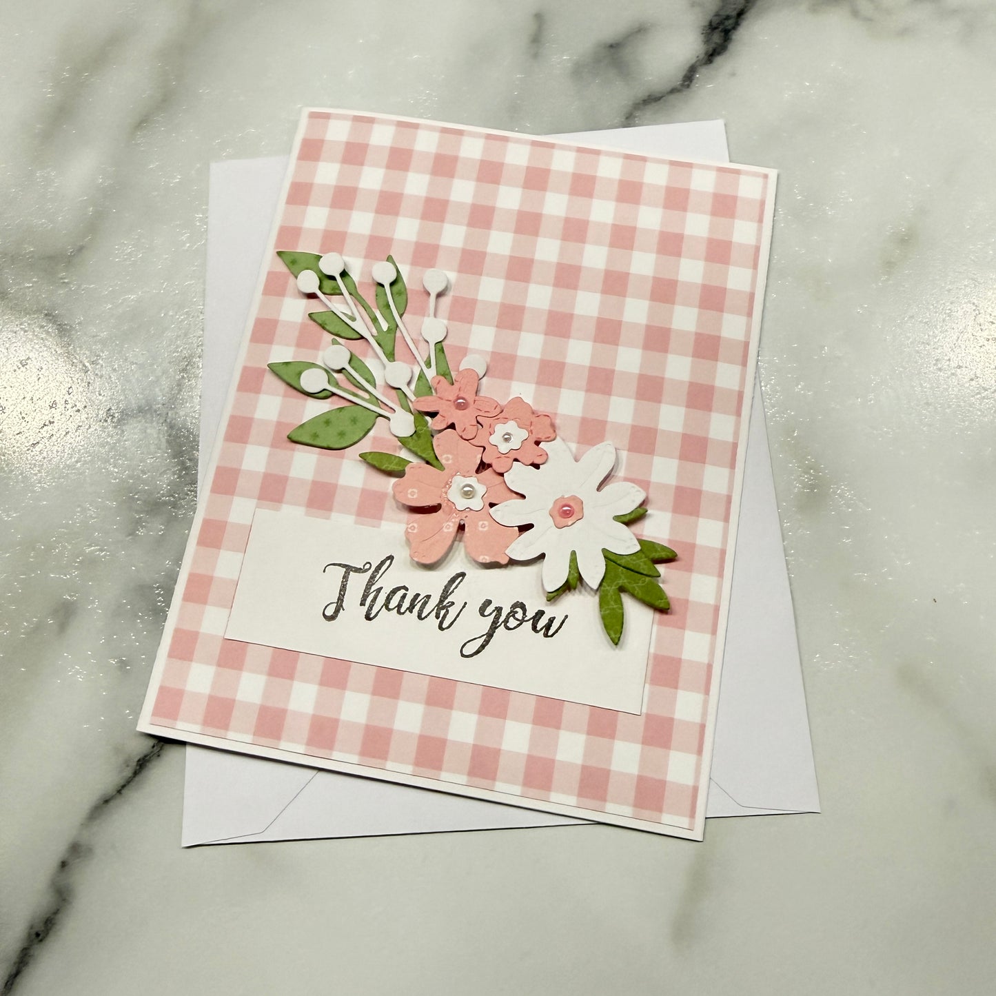 A handmade thank you card featuring a pink and white gingham pattern, layered paper flowers in pink, white, and green, and a white label with the words 'Thank you' in black script. The card is displayed on top of a white envelope on a light gray marble surface.