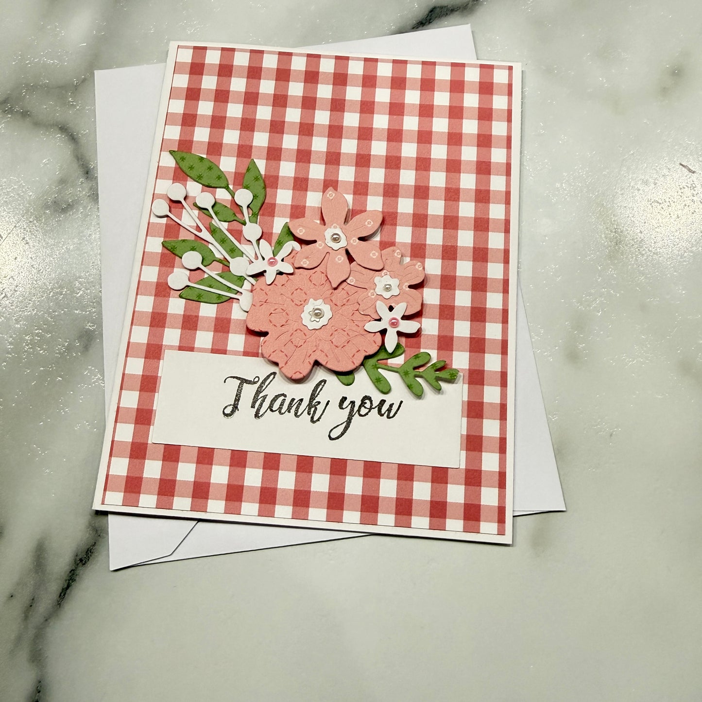 A handmade square thank you card with a red and white gingham pattern, featuring layered pink and white paper flowers with green leaves and the words 'Thank you' in black script. The card is displayed on top of a white envelope on a light gray marble surface.