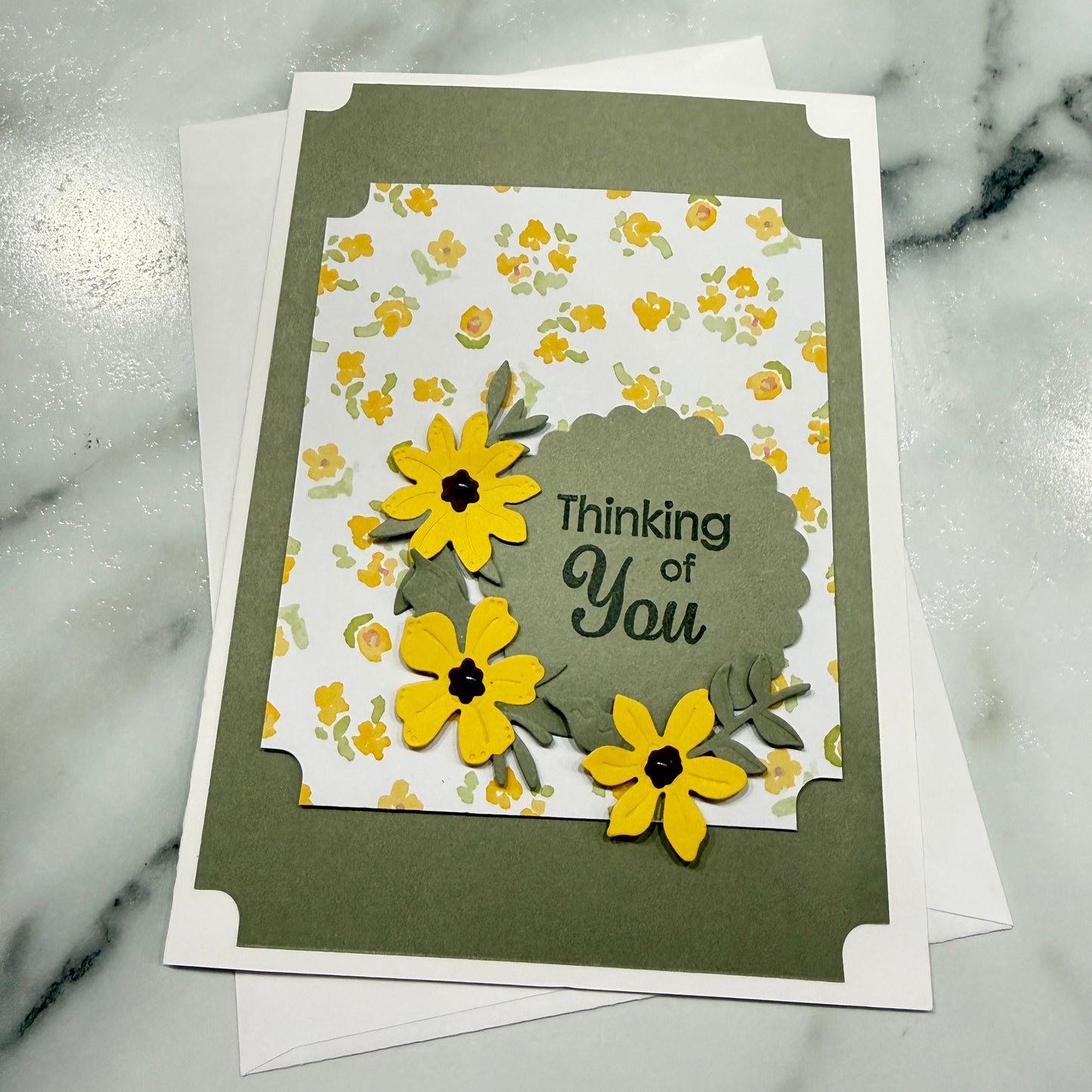 A handmade greeting card with a layered design, featuring yellow flower cutouts, green leaves, and the text 'Thinking of You' on a scalloped olive-green label, set against a white floral background and resting on a marble surface.