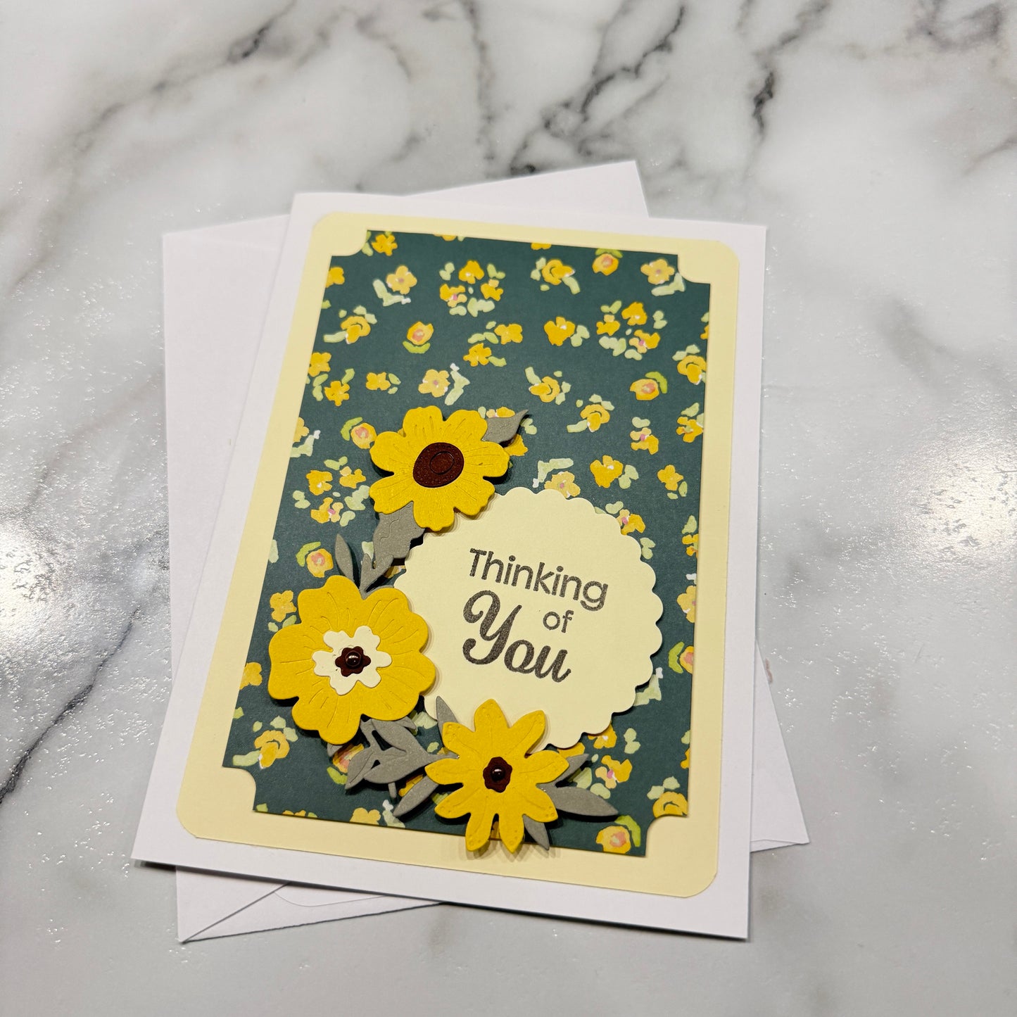 A handmade layered greeting card with a floral patterned background, yellow paper flowers, and a scalloped cream label reading 'Thinking of You,' photographed on a white marble surface with white envelopes behind it.