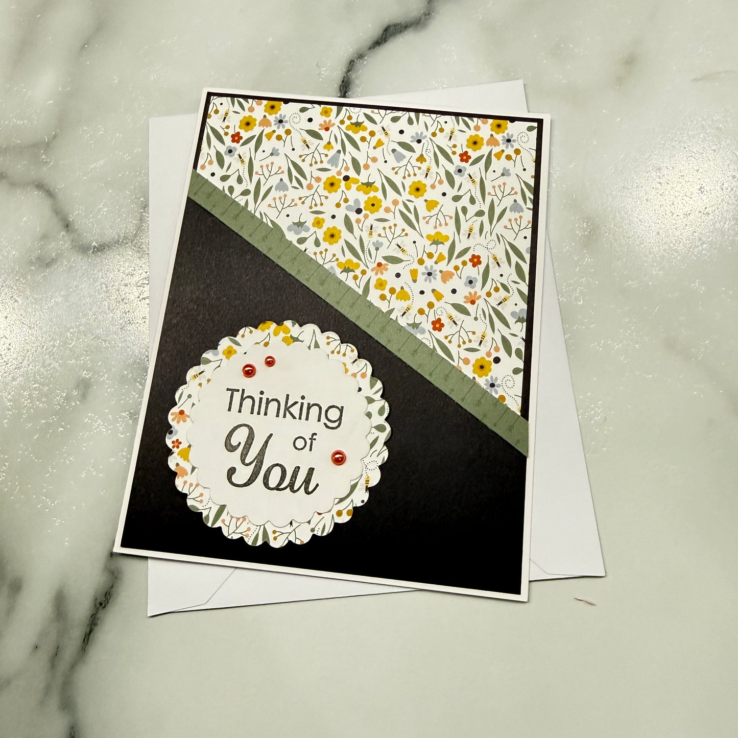 A handmade greeting card with a black background, a diagonal olive green stripe, a floral patterned top section, and a scalloped white circle reading 'Thinking of You,' placed on a white envelope over a light marble surface.