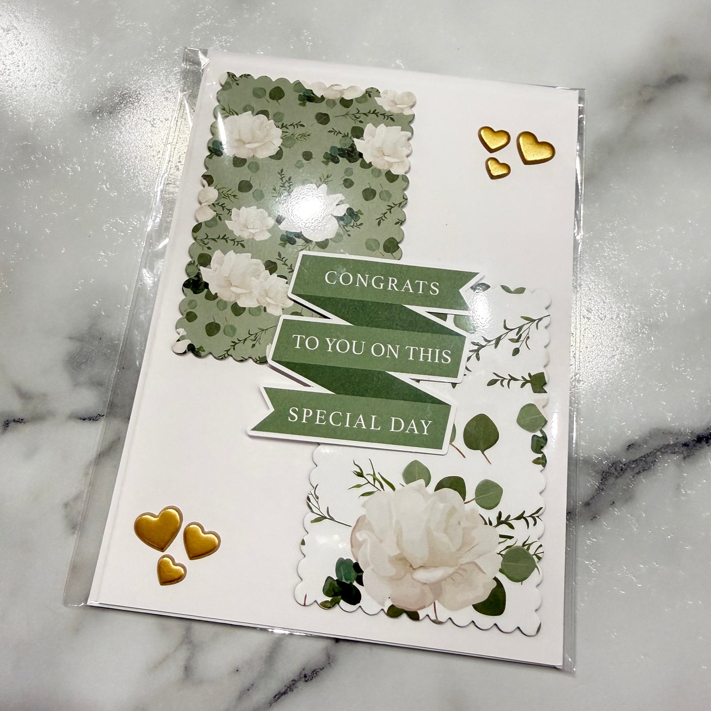 A handmade congratulations card in clear plastic packaging, featuring green and white floral designs, scalloped borders, and gold heart stickers on a white marble background.