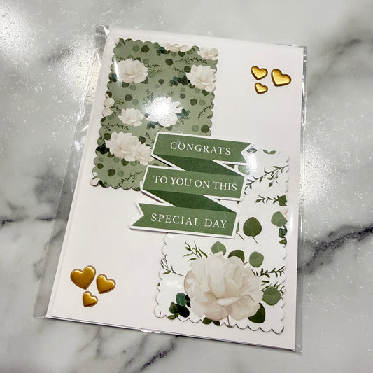 A handmade congratulations card in clear plastic packaging, featuring green and white floral designs, scalloped borders, and gold heart stickers on a white marble background.