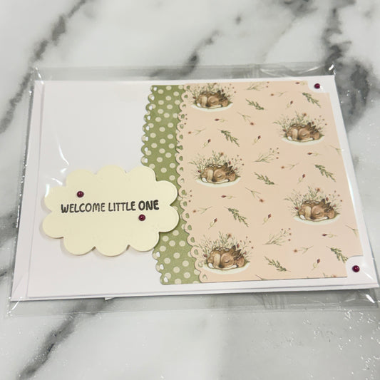 A handmade baby shower card in clear plastic packaging, featuring a cream scalloped tag that reads 'WELCOME LITTLE ONE' and a patterned panel with deer sleeping.