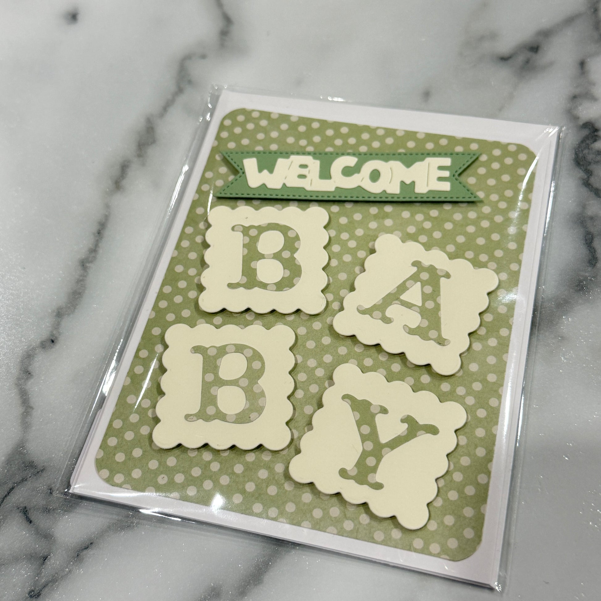 A packaged baby shower card featuring a green and white polka dot background with cream-colored scalloped letters spelling 'BABY' and a green banner that reads 'WELCOME'. The card is sealed in clear plastic packaging and photographed on a white and gray marble surface.