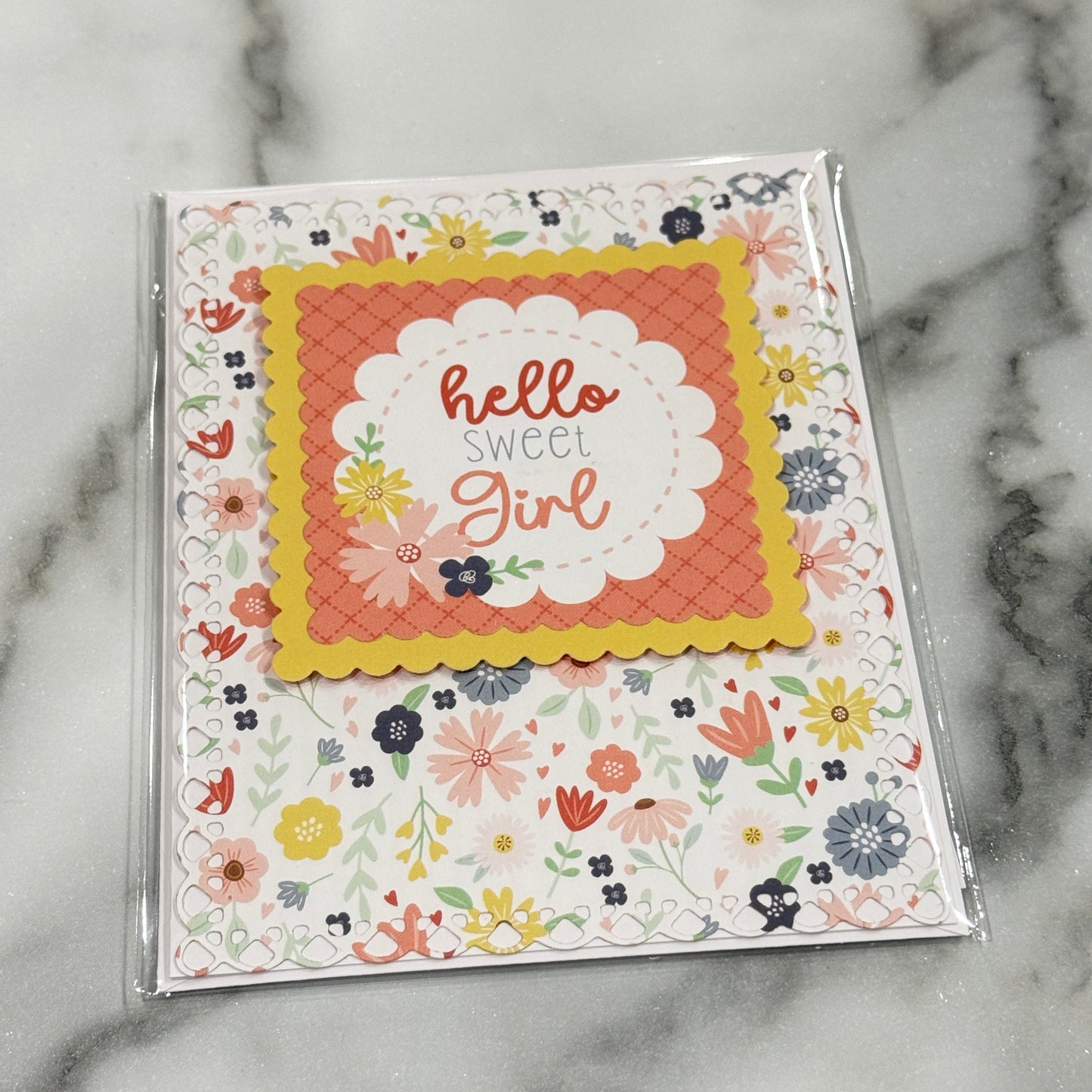 A handmade greeting card in clear plastic packaging, featuring a central scalloped white label with the words 'hello sweet girl' in coral and gray script, surrounded by a coral-orange patterned square and a yellow border. The card is mounted on a white background with a multicolor floral print, set on a gray and white marble surface.