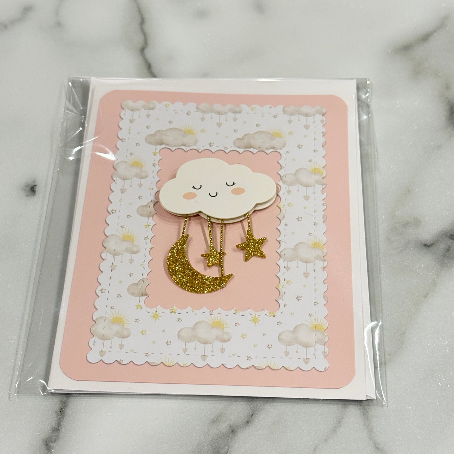 A baby shower themed card featuring a cute cloud and moon design, sealed in a clear plastic sleeve and placed on a white marble surface.