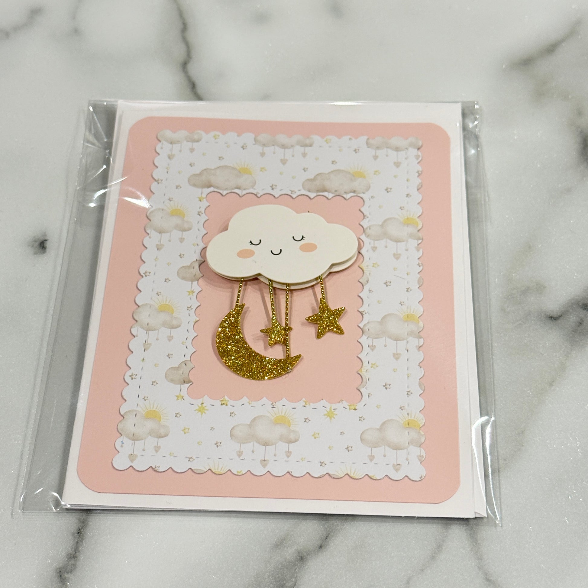 A baby shower themed card featuring a cute cloud and moon design, sealed in a clear plastic sleeve and placed on a white marble surface.