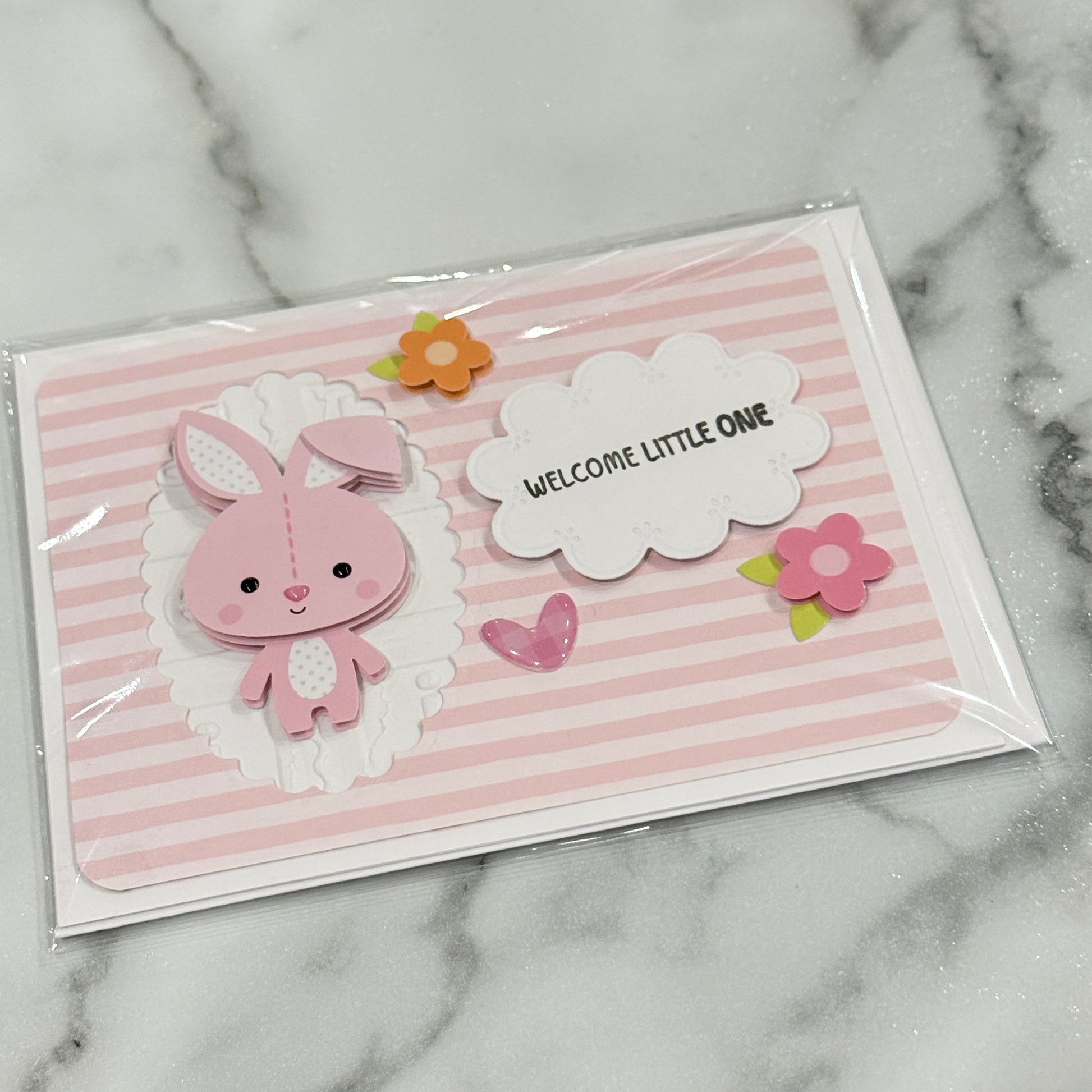 A handmade baby shower card featuring a pink bunny, pink and white stripes, and a white cloud that reads 'WELCOME LITTLE ONE', sealed in a clear plastic sleeve and placed on a white marble surface.