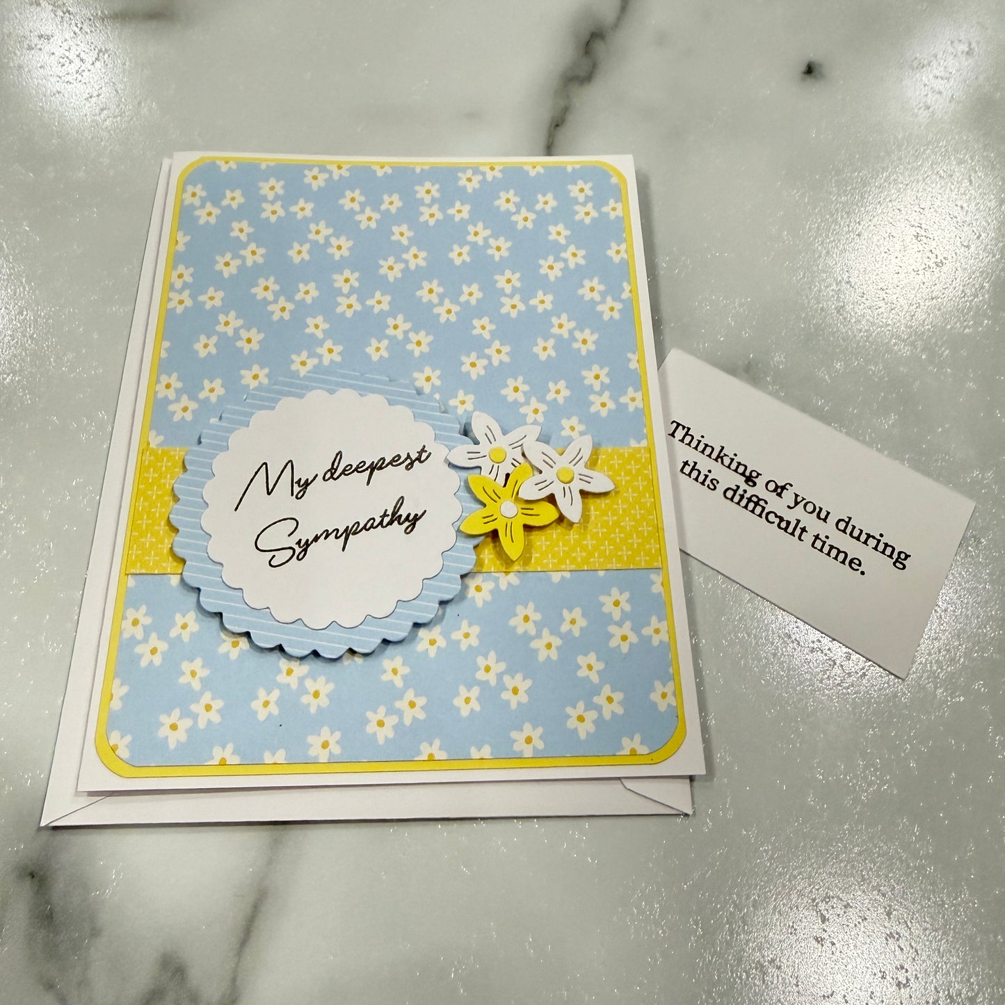 A handmade sympathy card with a light blue and yellow daisy pattern, a scalloped white label reading 'My deepest Sympathy,' and two small layered flowers, shown next to a small note on a marble surface.