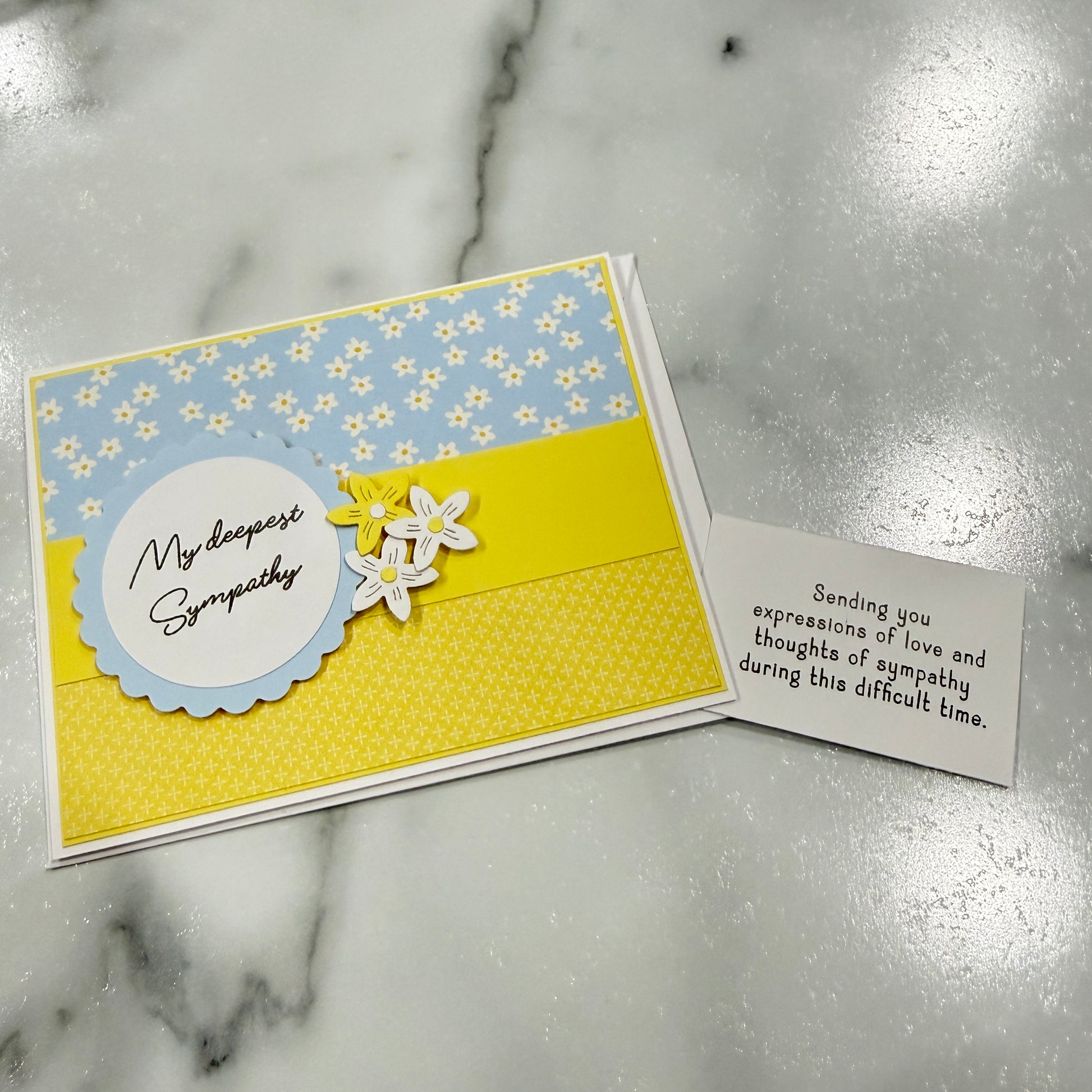 A handmade sympathy greeting card with a light blue and yellow floral patterned top section, a pale blue scalloped circle containing the words 'My deepest Sympathy' in black script, and yellow and white flower embellishments. A separate small white card with a sympathy message is placed beside it on a white marble surface.