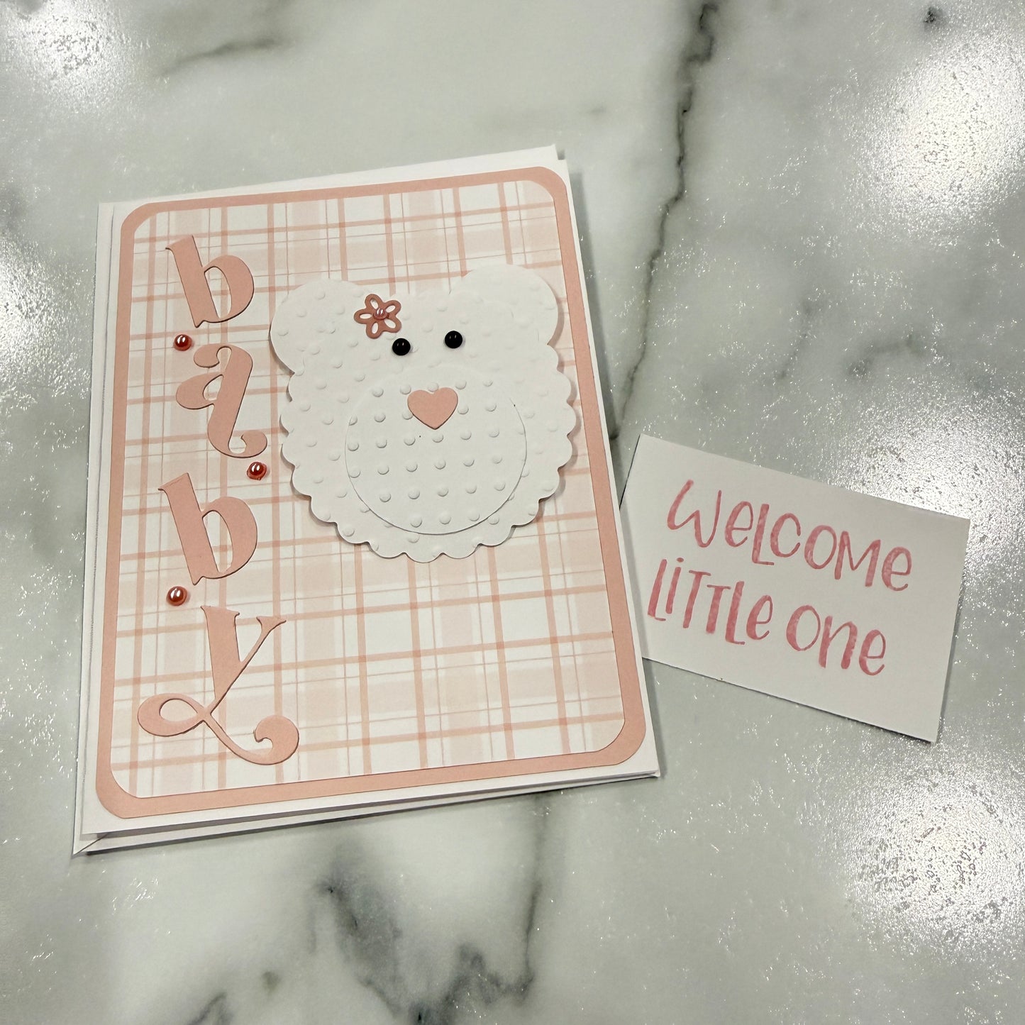 A handmade baby shower card set featuring a square card with a pink and white plaid background, a white bear face with a small flower on its head, and the word 'baby' in decorative script, next to a small white card that reads 'Welcome little one' on a marble surface.