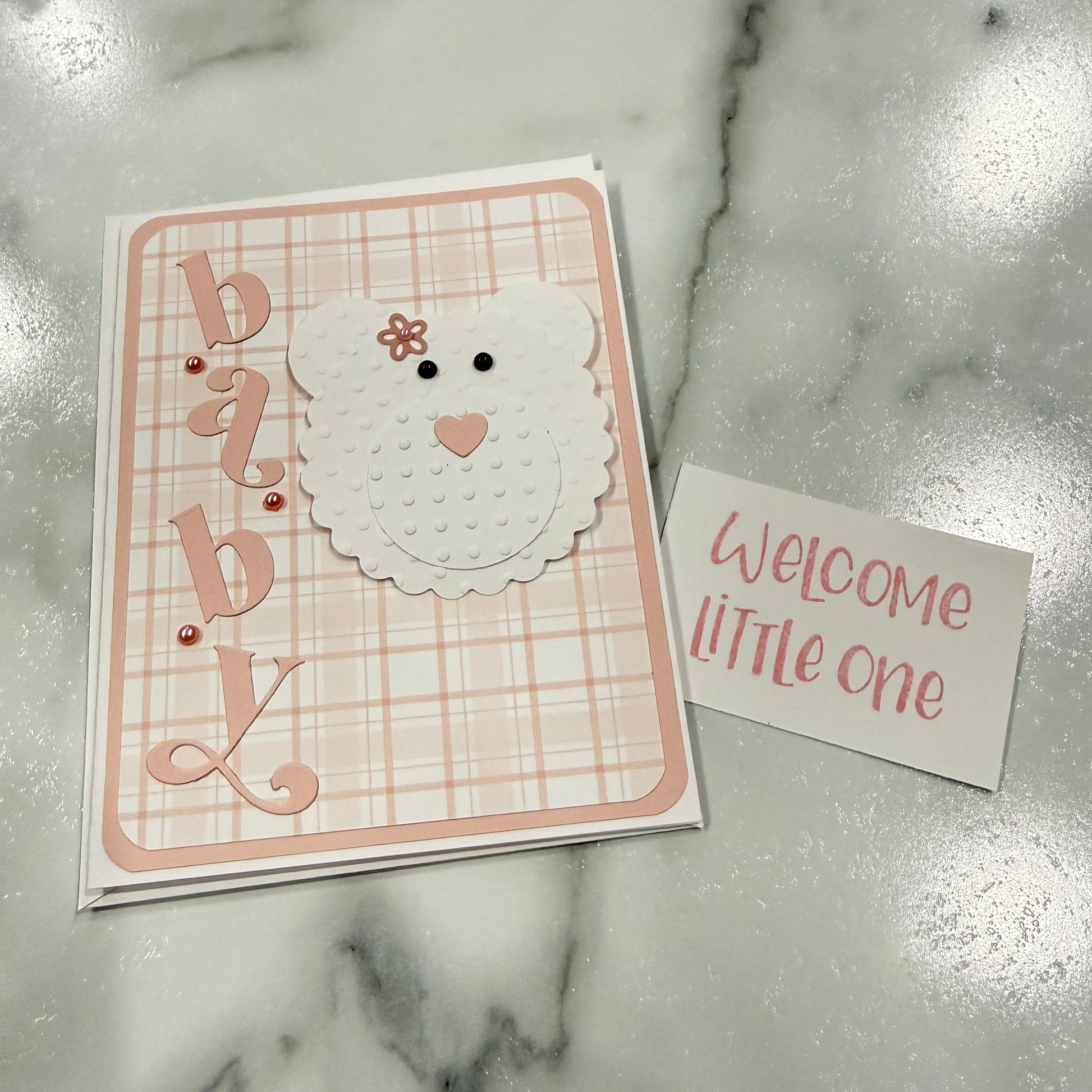 A handmade baby shower card set featuring a square card with a pink and white plaid background, a white bear face with a small flower on its head, and the word 'baby' in decorative script, next to a small white card that reads 'Welcome little one' on a marble surface.