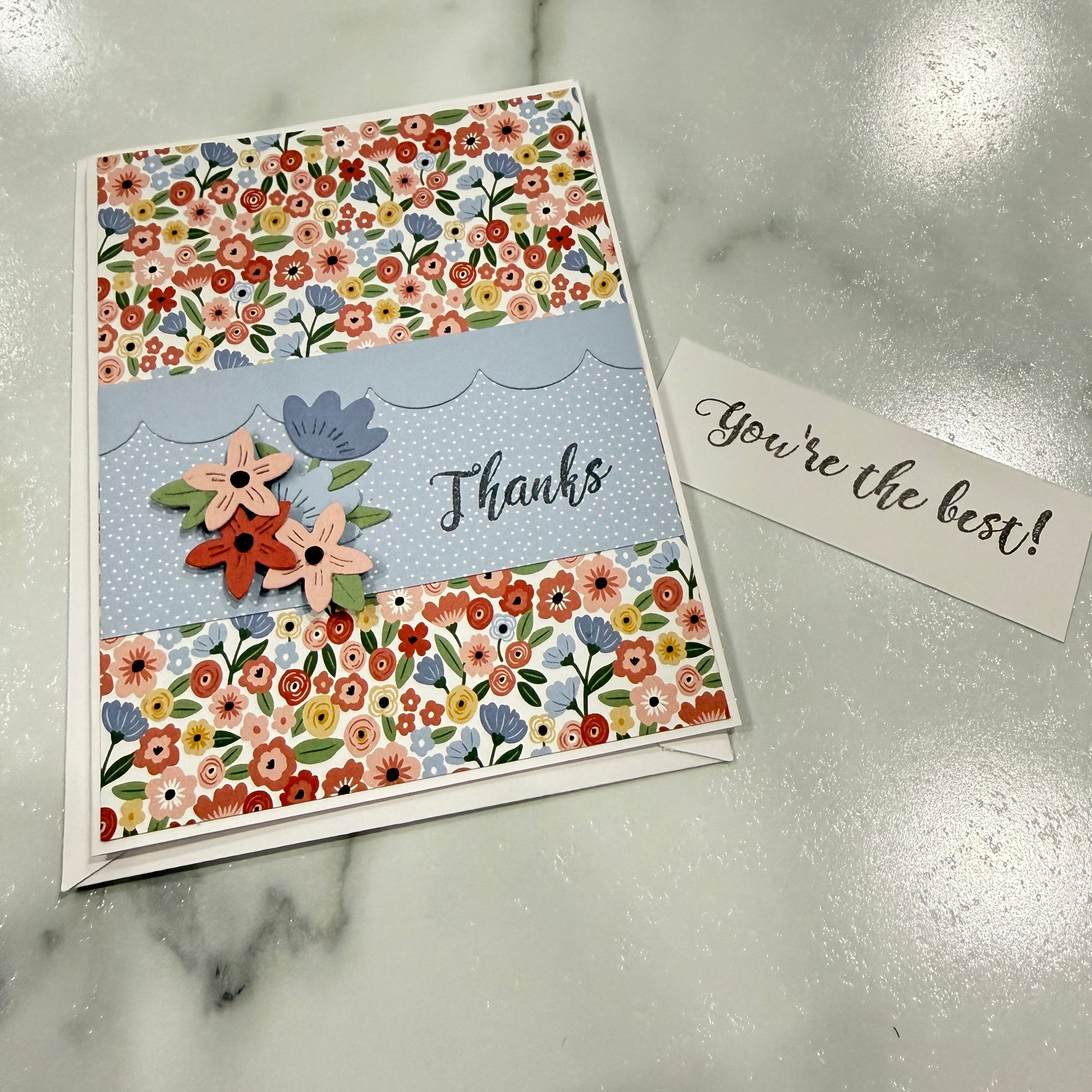 A handmade floral thank you card with a matching small note card on a light gray marble surface. The card features a light blue center band with the word 'Thanks' and three layered paper flowers, framed by two floral patterned borders at the top and bottom.