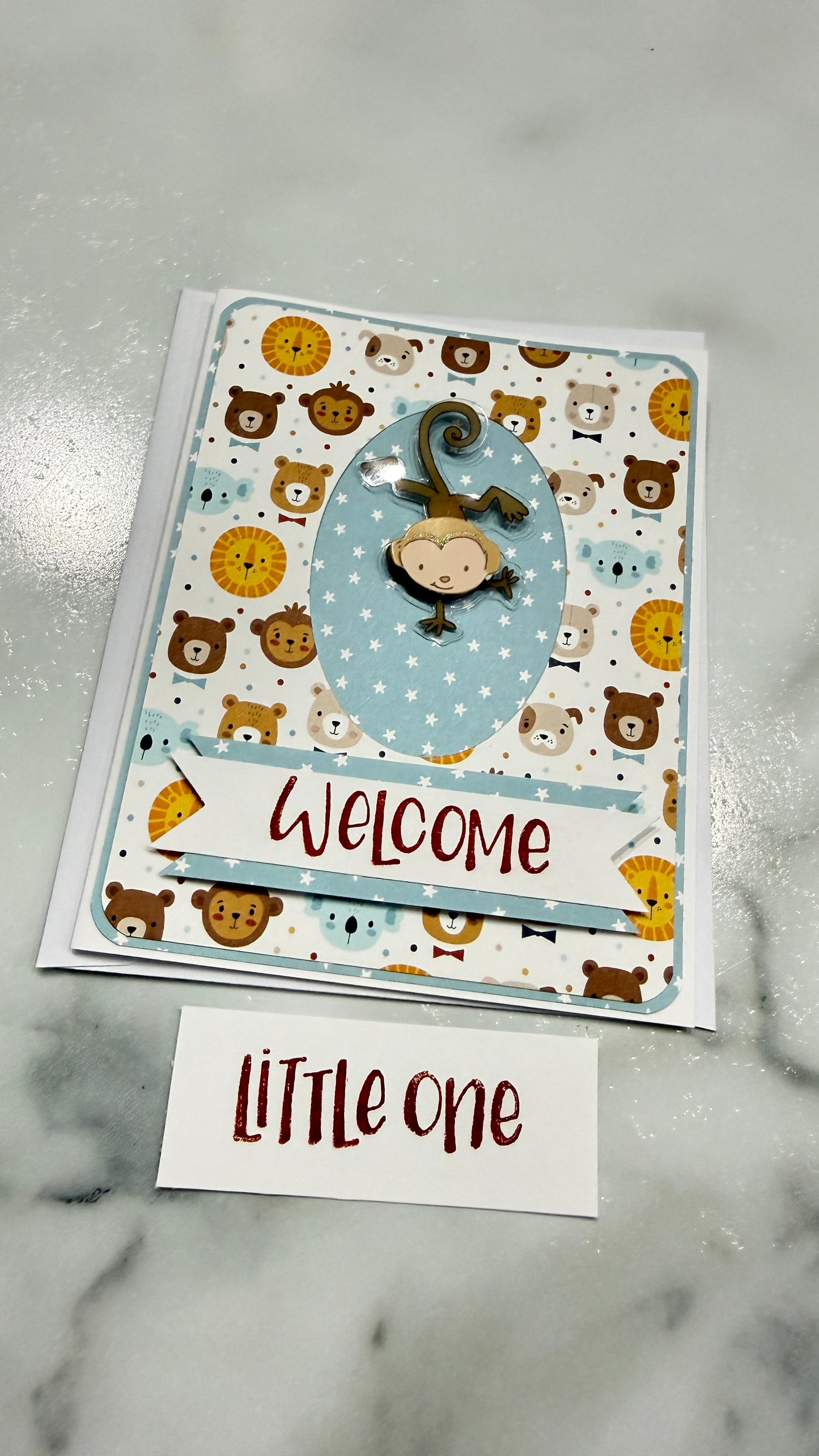 A handmade baby welcome card featuring a playful pattern of cartoon bears and monkeys on a white background, with a central blue oval and a 'Welcome' banner. A small white card below reads 'LITTLE one' in red script, all placed on a light gray marble surface.