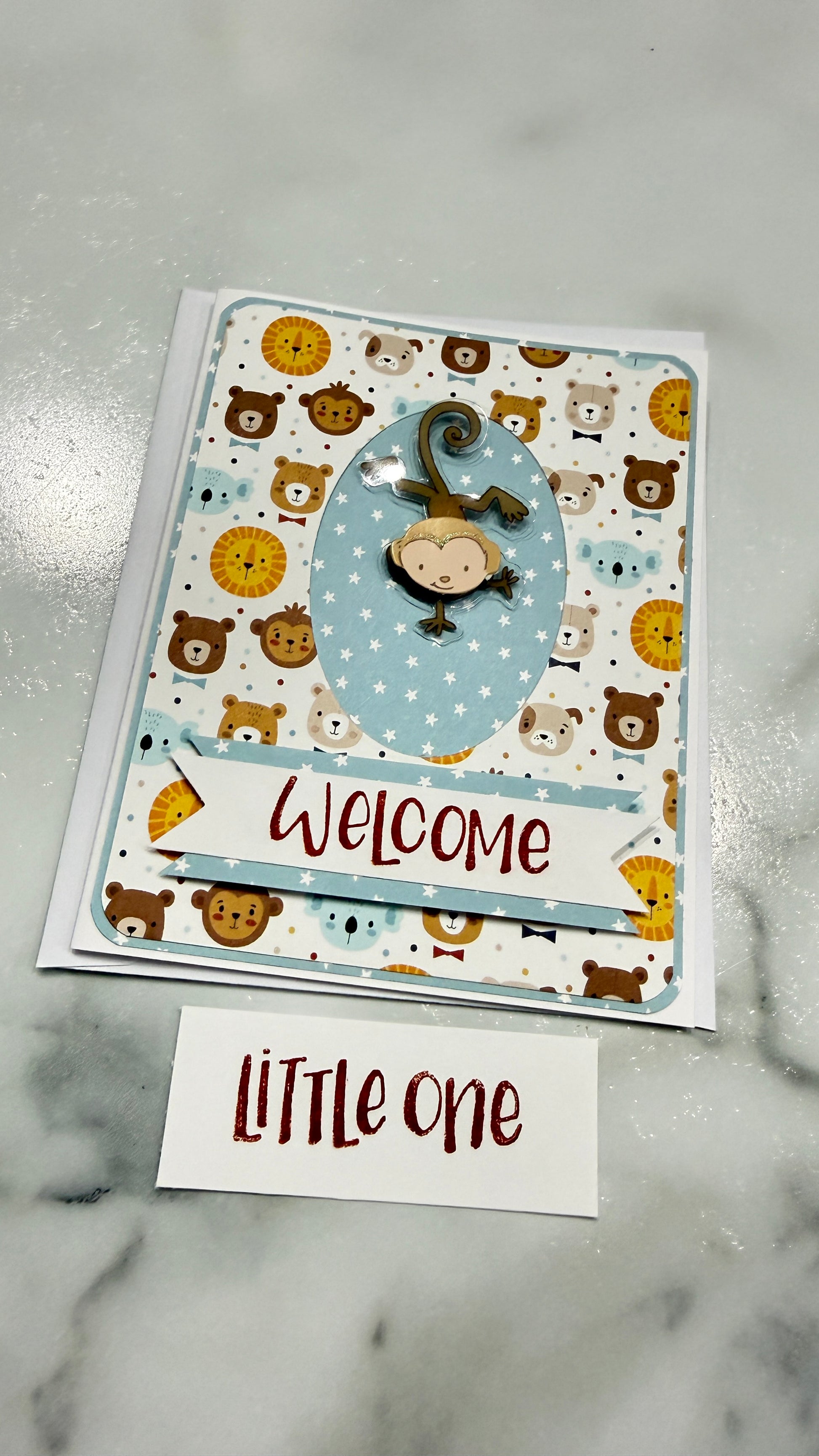 A handmade baby welcome card featuring a playful pattern of cartoon bears and monkeys on a white background, with a central blue oval and a 'Welcome' banner. A small white card below reads 'LITTLE one' in red script, all placed on a light gray marble surface.