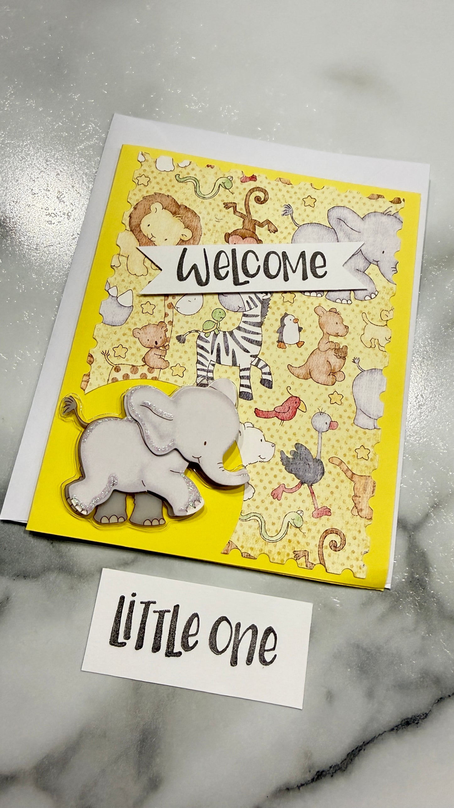 A handmade safari-themed greeting card with a yellow background, cartoon animal illustrations, a white elephant die-cut layer, and a white 'Welcome' banner, shown on a marble surface with a small card reading 'Little one'.