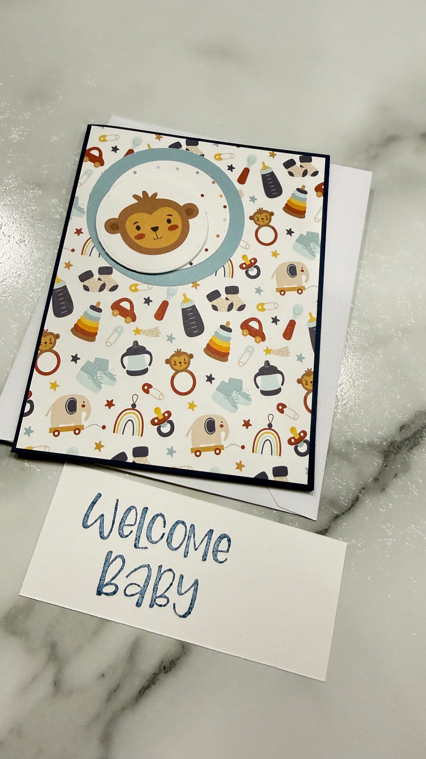 A handmade baby welcome card set featuring a square patterned card with a cartoon monkey and baby items, a white envelope, and a separate white card that says 'Welcome Baby' in blue script, placed on a gray marble surface.