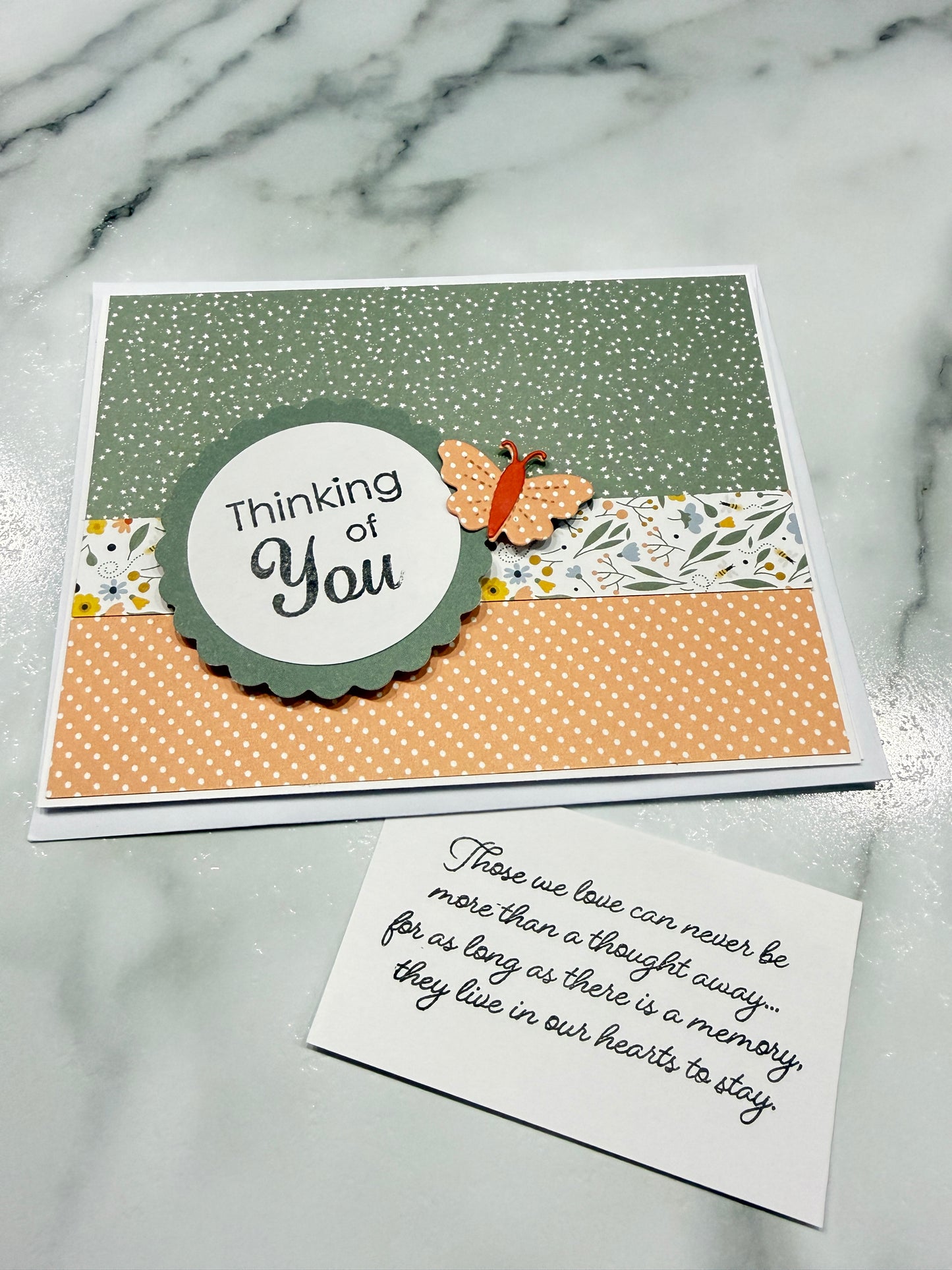 A handmade greeting card with layered paper designs and a butterfly embellishment, shown next to a small handwritten note on a light marble surface.