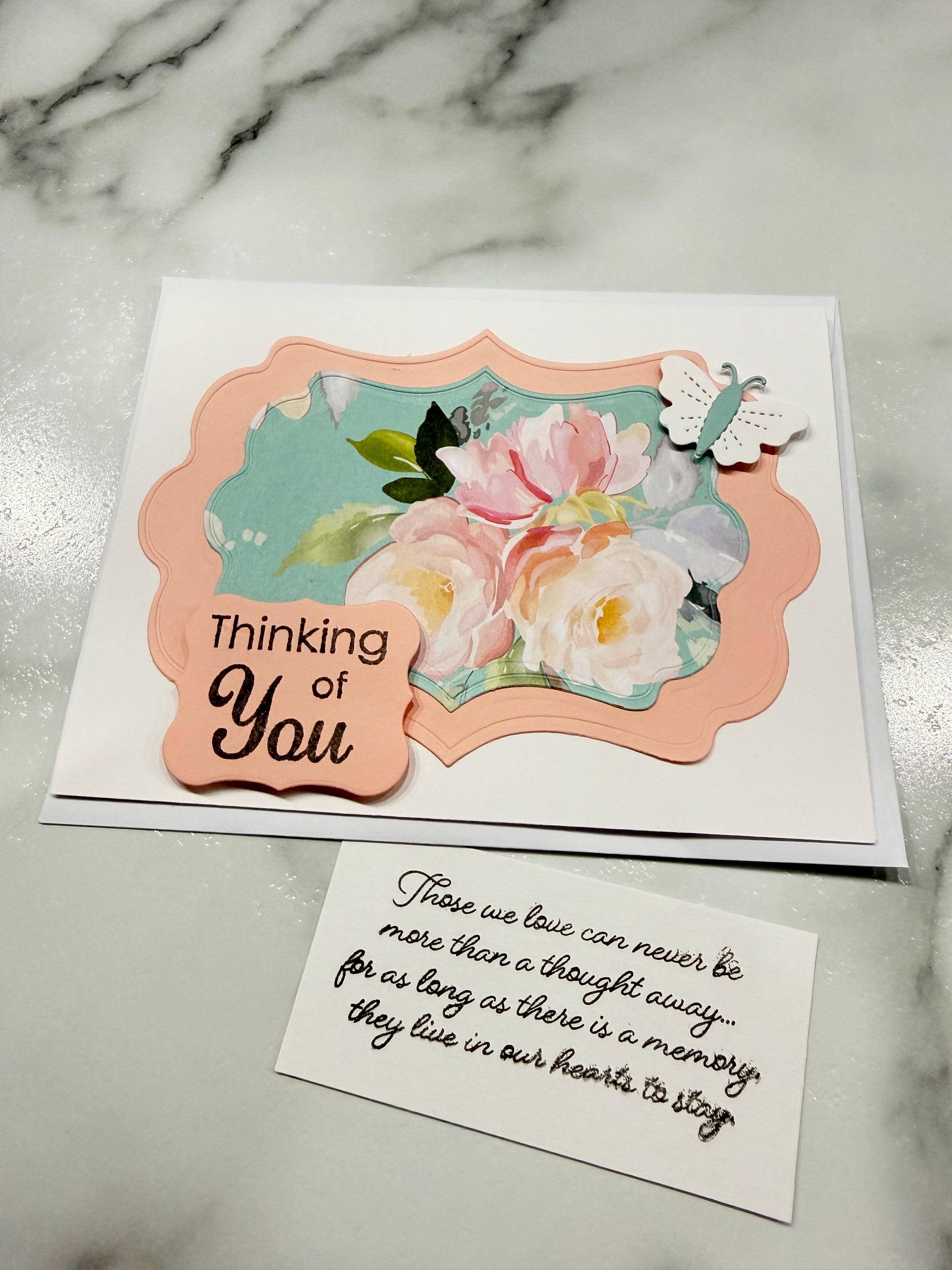A handmade sympathy card with a white base, a peach scalloped frame, pastel pink and white roses, green leaves, and a small peach tag reading 'Thinking of You,' shown beside a small white card with handwritten text on a light marble surface.