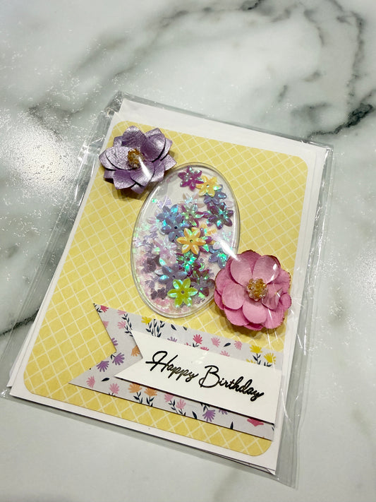 A handmade birthday card in clear plastic packaging, featuring a yellow patterned background, an oval clear resin compartment filled with colorful glitter, pink and purple paper flowers, and a white banner that reads 'Happy Birthday'.