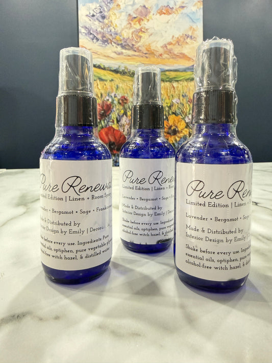 Three blue glass spray bottles with white labels reading 'Pure Renewal' are arranged on a white marble surface next to a dark gray background and a colorful painting.