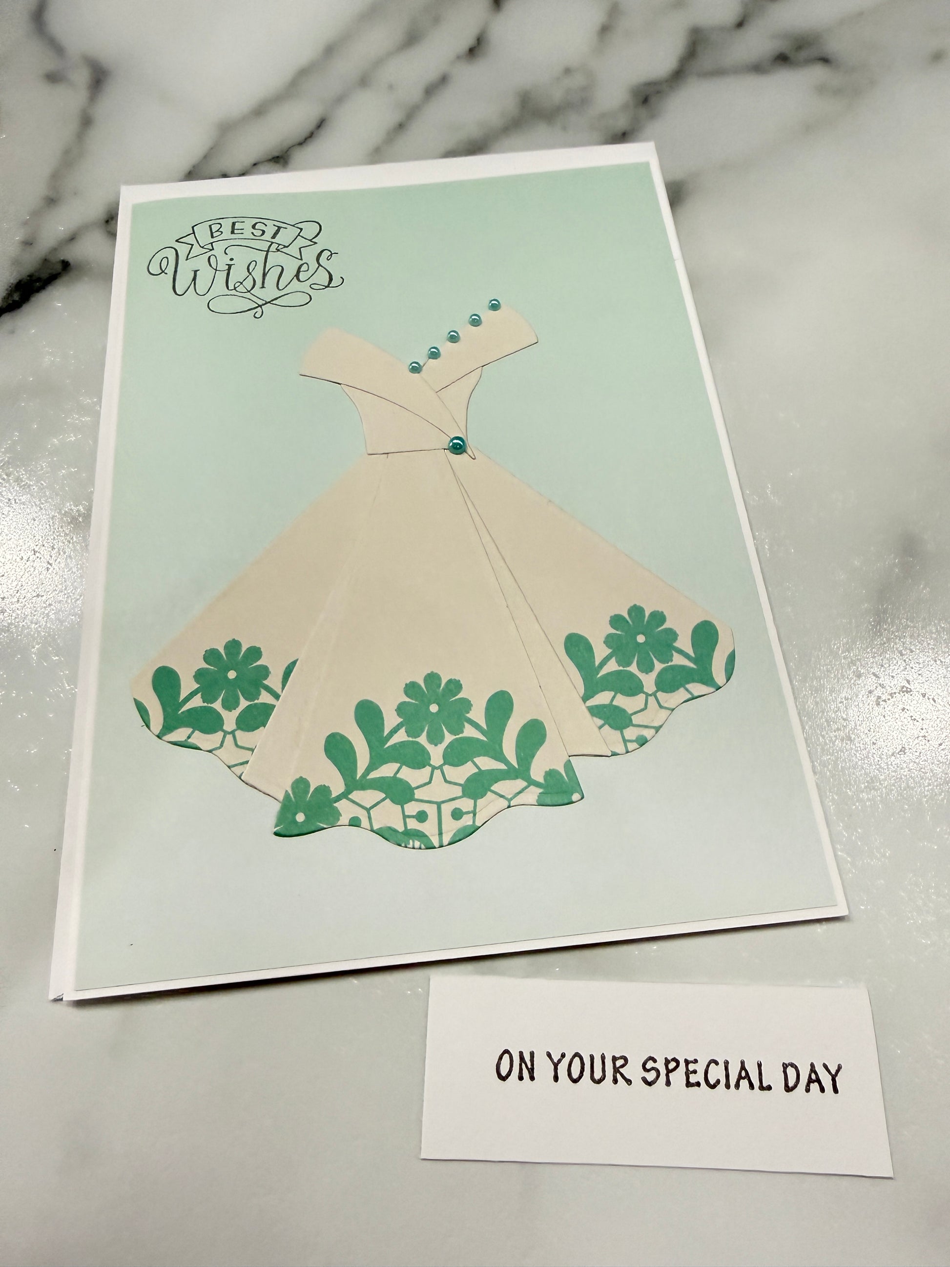 A square greeting card featuring a cream-colored dress with green floral patterns on a pale mint background, placed on a white marble surface next to a small white card that reads 'ON YOUR SPECIAL DAY'.