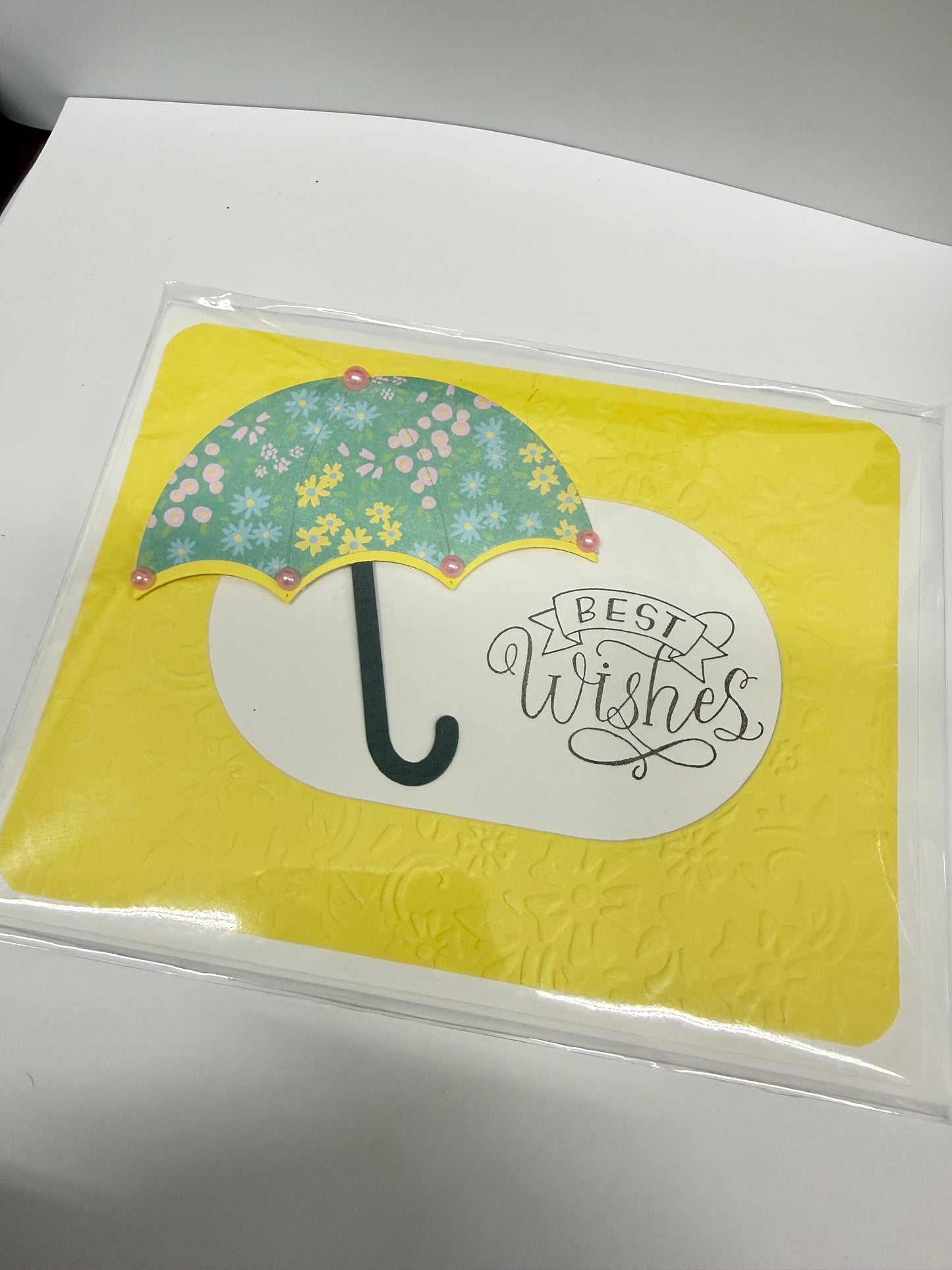 A square yellow card or coaster inside a clear plastic sleeve, featuring a white oval with the phrase 'BEST WISHES' in script and a floral green umbrella illustration on a white surface.