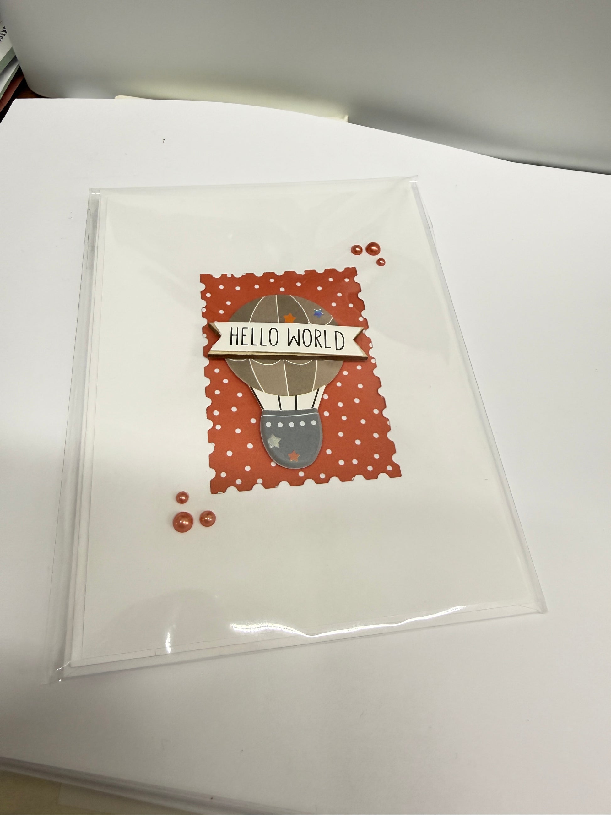 A clear plastic sleeve containing a square greeting card with a hot air balloon illustration and the words 'HELLO WORLD' on a white background.