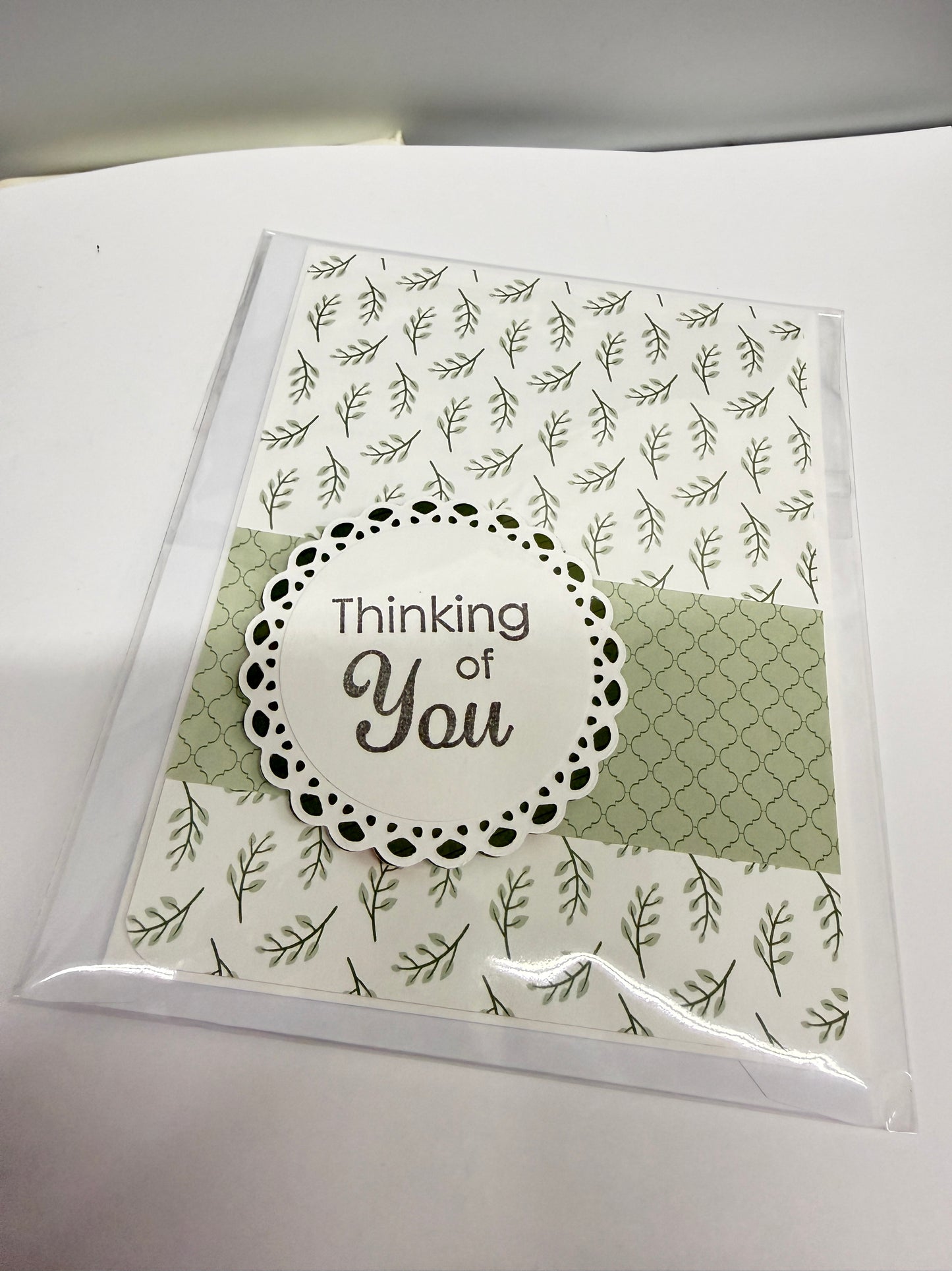 A handmade greeting card in clear plastic packaging, featuring a white background with green leaf patterns, a sage green patterned panel, and a central white circular label with the words 'Thinking of You'.