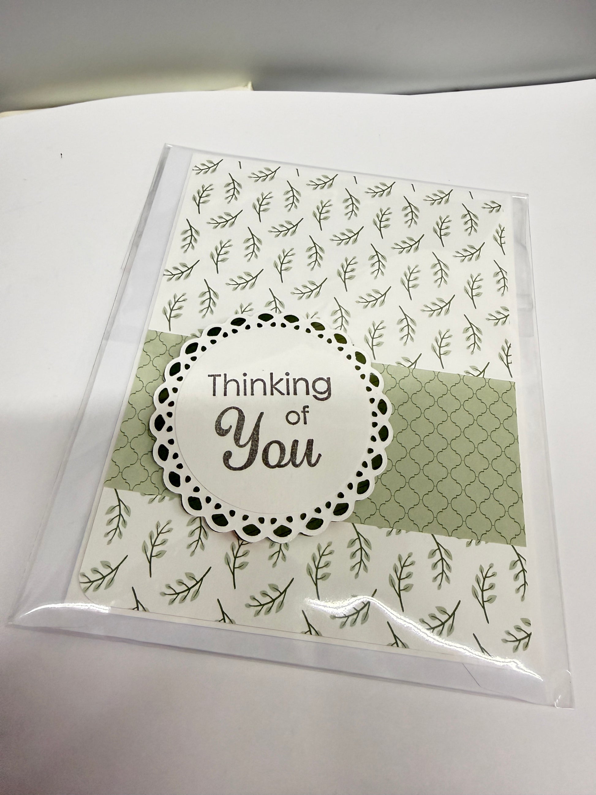 A handmade greeting card in clear plastic packaging, featuring a white background with green leaf patterns, a sage green patterned panel, and a central white circular label with the words 'Thinking of You'.