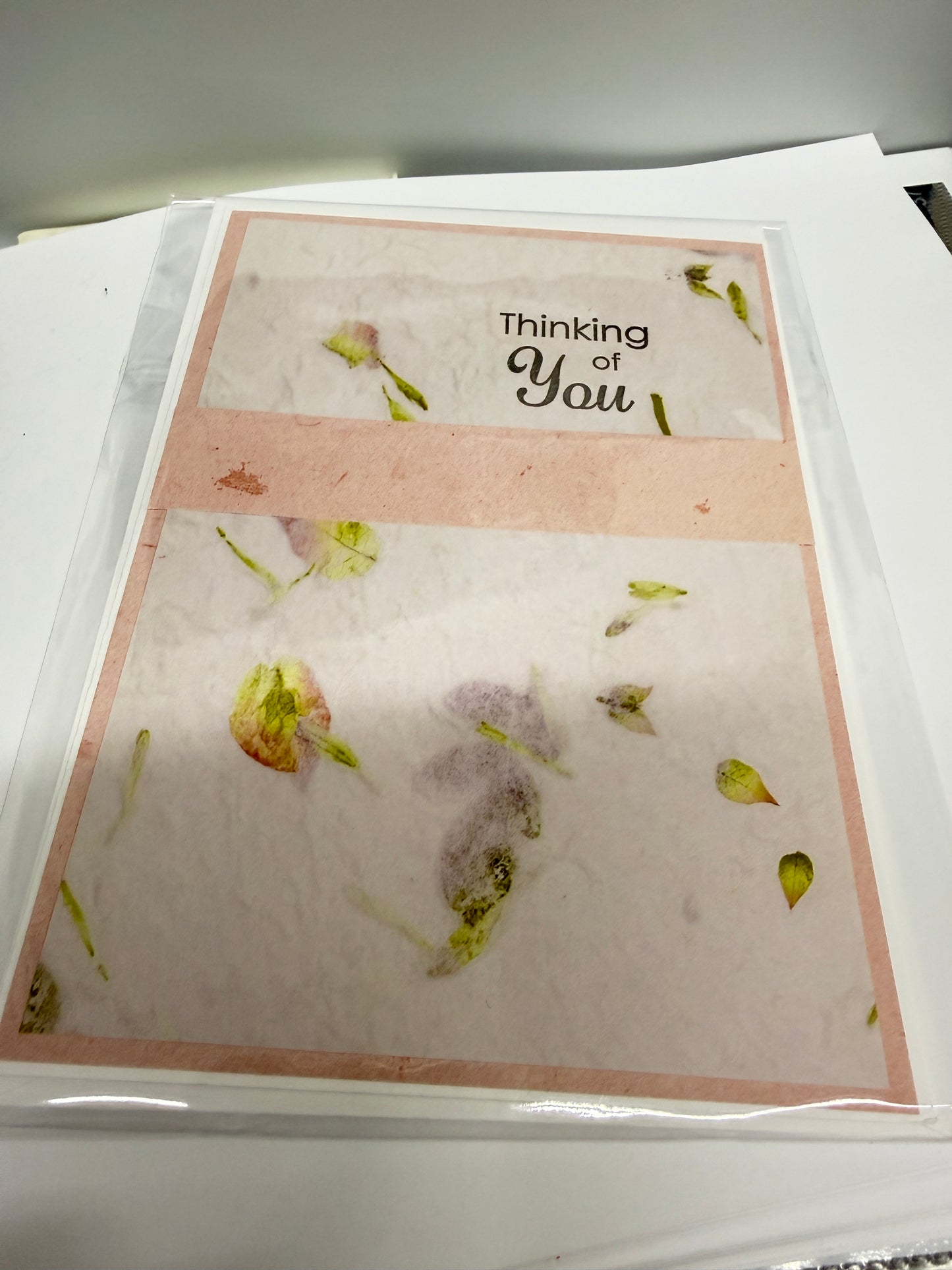 A folded greeting card with a pink border and scattered pressed flower petals, placed inside a clear plastic sleeve on a white surface. The left panel reads 'Thinking of You'.