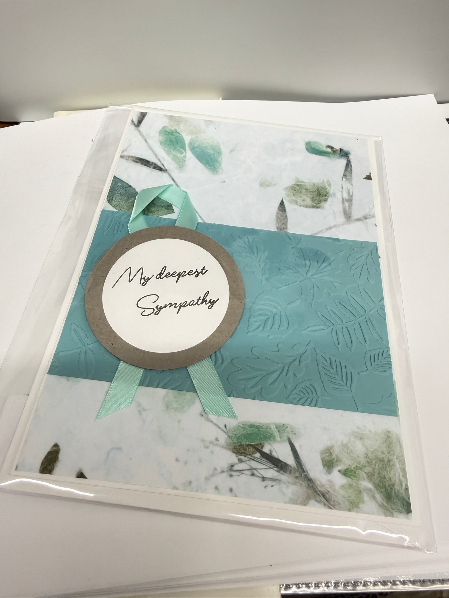 A handmade greeting card in a clear plastic sleeve, featuring a central teal textured panel, white side panels with green botanical illustrations, a light green ribbon, and a round brown and white label that reads 'My Deepest Sympathy'.