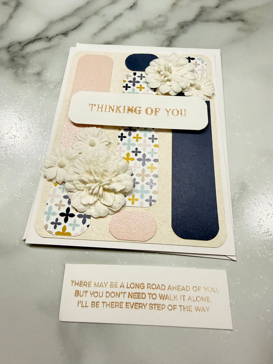 A handmade layered greeting card with floral embellishments and a vertical tab reading 'THINKING OF YOU,' shown next to a small card with a message on a marble surface.
