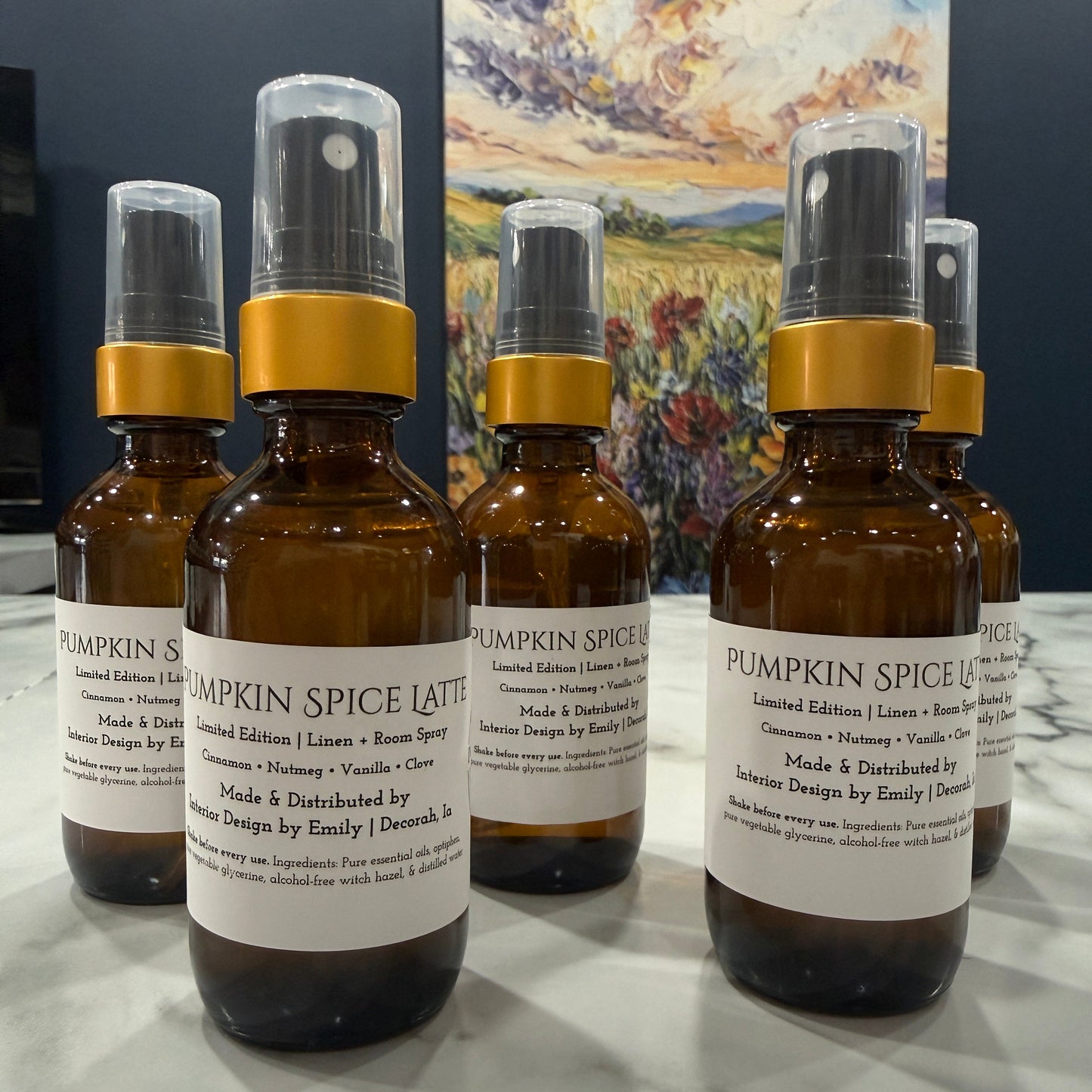 Five amber glass spray bottles with white labels and gold collars arranged on a light marble surface, with a dark wall and colorful landscape painting in the background.