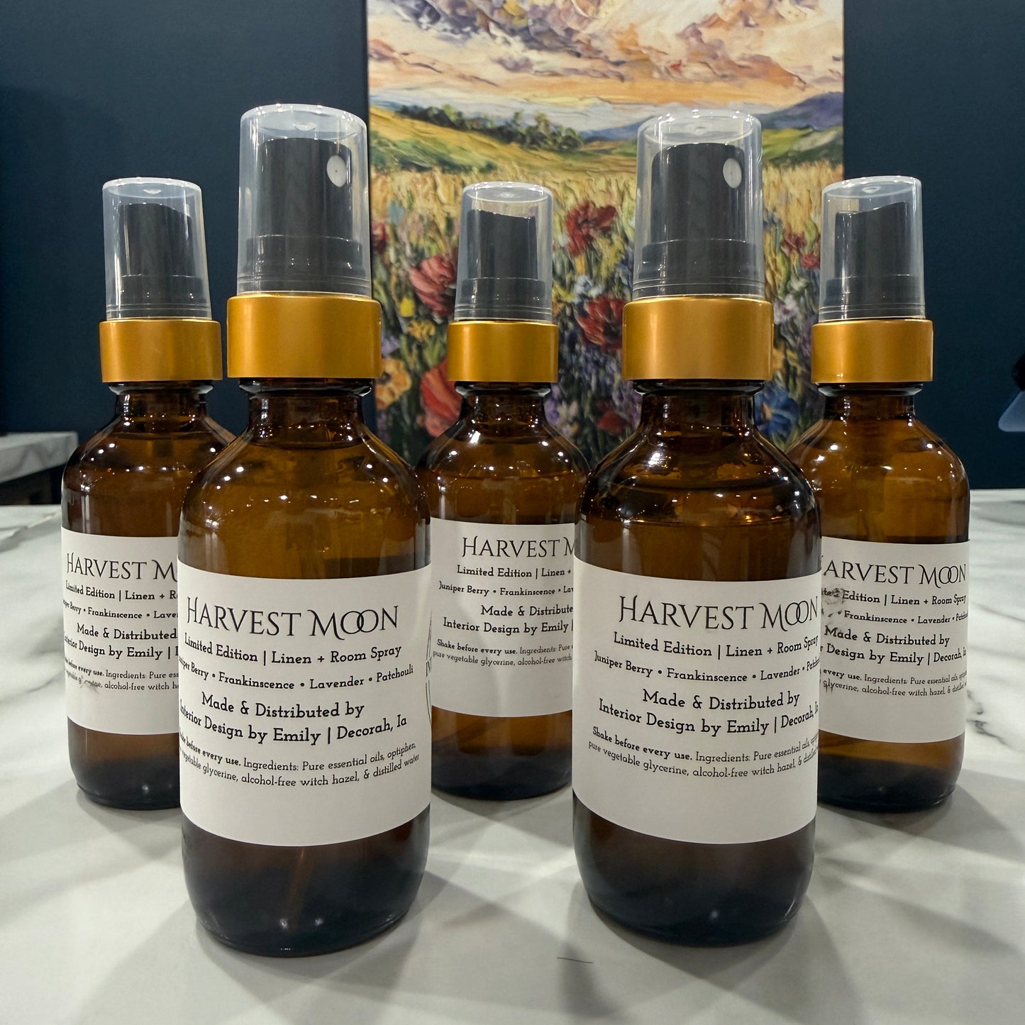 Five amber glass spray bottles with gold collars and clear spray tops arranged on a white surface, with visible Harvest Moon labels. A colorful landscape painting hangs on the wall behind them.