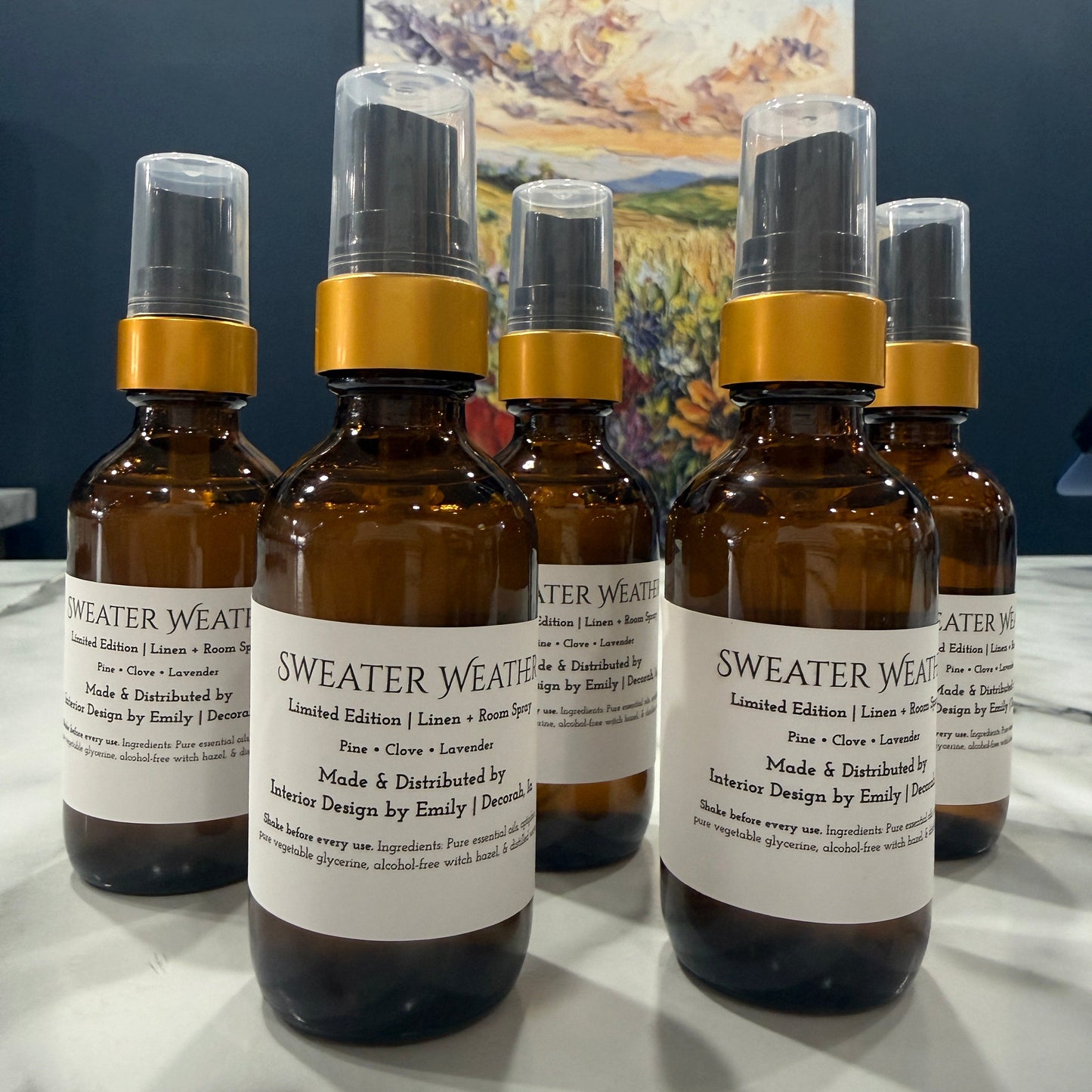 Five amber glass spray bottles labeled 'SWEATER WEATHER' with gold collars and clear caps, arranged on a white marble surface with a dark blue wall and a mountain painting in the background.