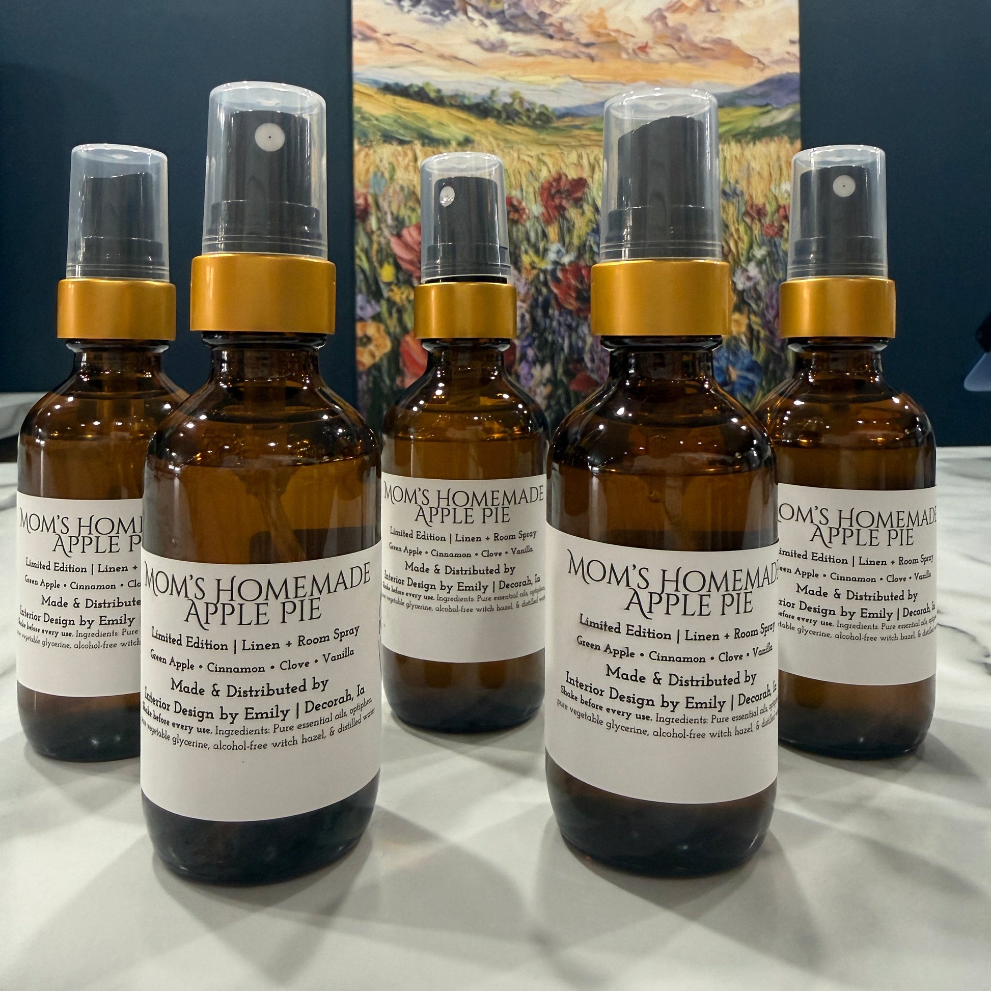 Five amber glass spray bottles labeled 'Mom's Homemade Apple Pie' with gold collars and clear caps, arranged on a white surface in front of a colorful landscape painting.