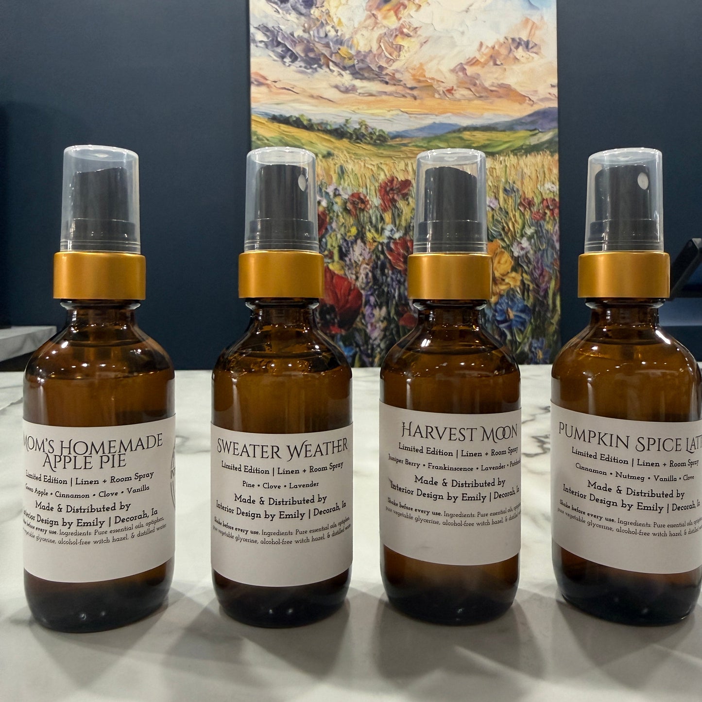 Four amber glass spray bottles with white labels and gold collars arranged on a white surface, with a colorful landscape painting hanging on the wall behind them.
