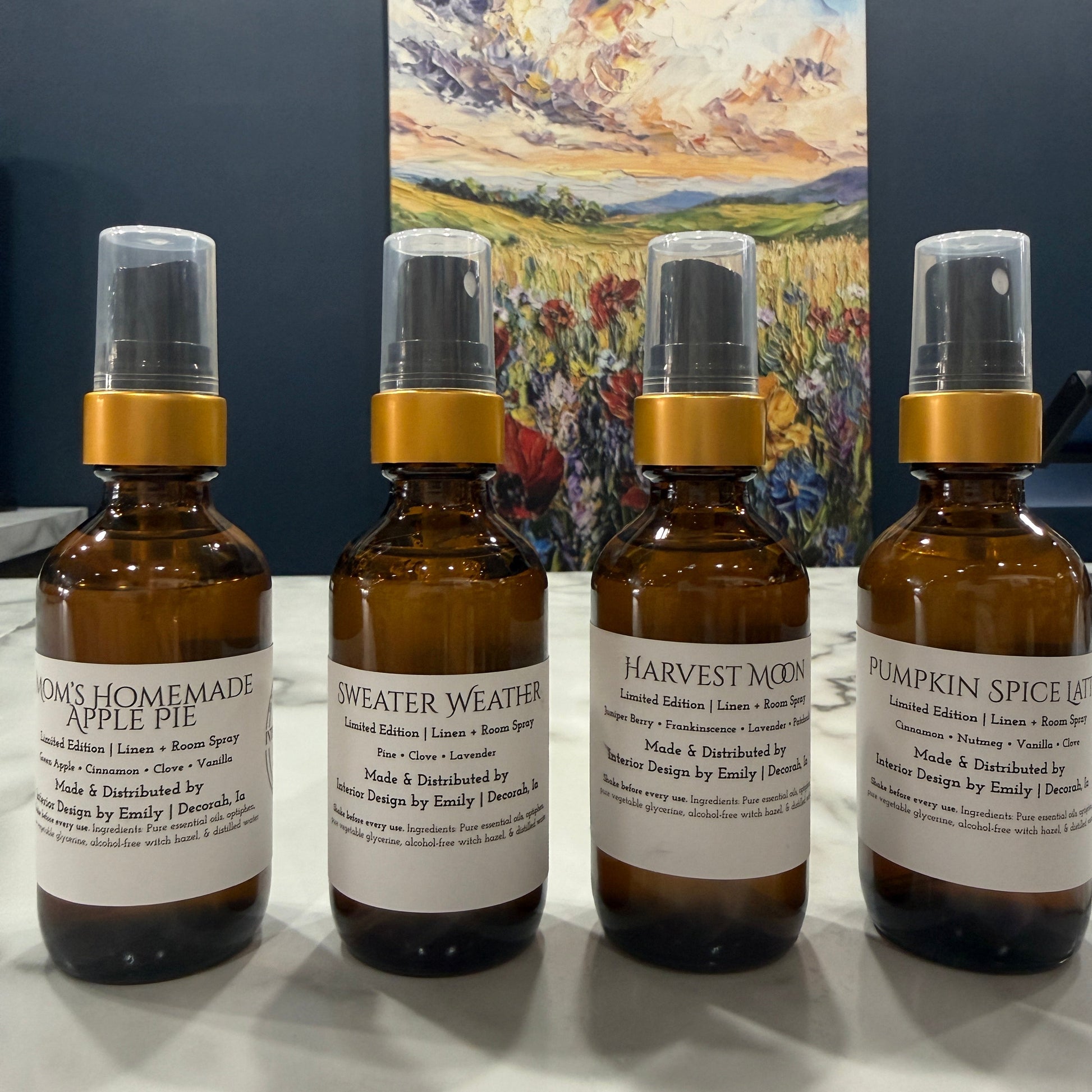 Four amber glass spray bottles with white labels and gold collars arranged on a white surface, with a colorful landscape painting hanging on the wall behind them.