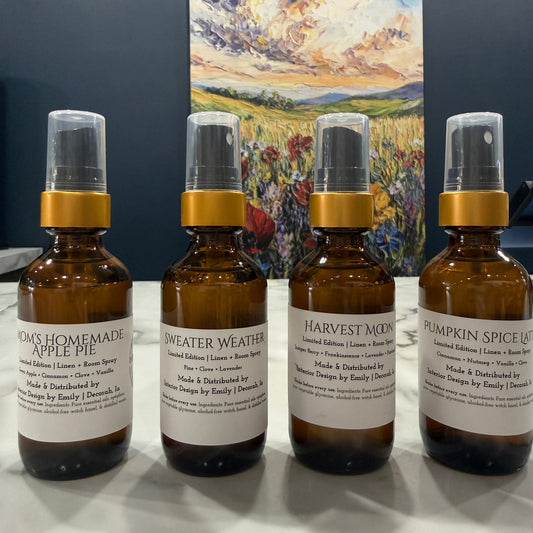 Four amber glass spray bottles with white labels and gold collars arranged on a white surface, with a colorful landscape painting hanging on the wall behind them.