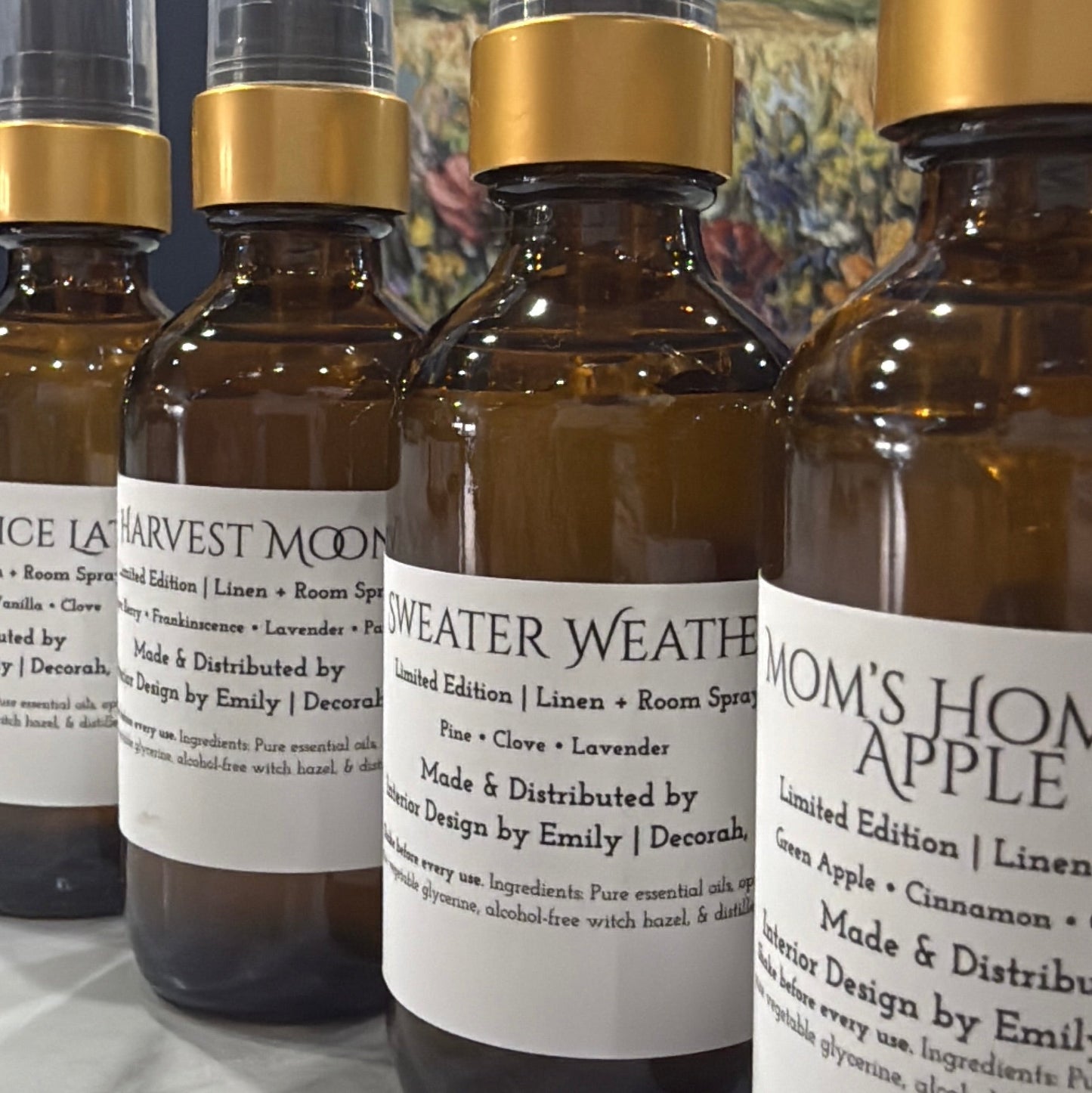Four amber glass spray bottles with black atomizers and gold collars, lined up on a marble surface in front of a colorful landscape painting. The front bottle has a white label reading 'MOM'S HOME APPLE'.