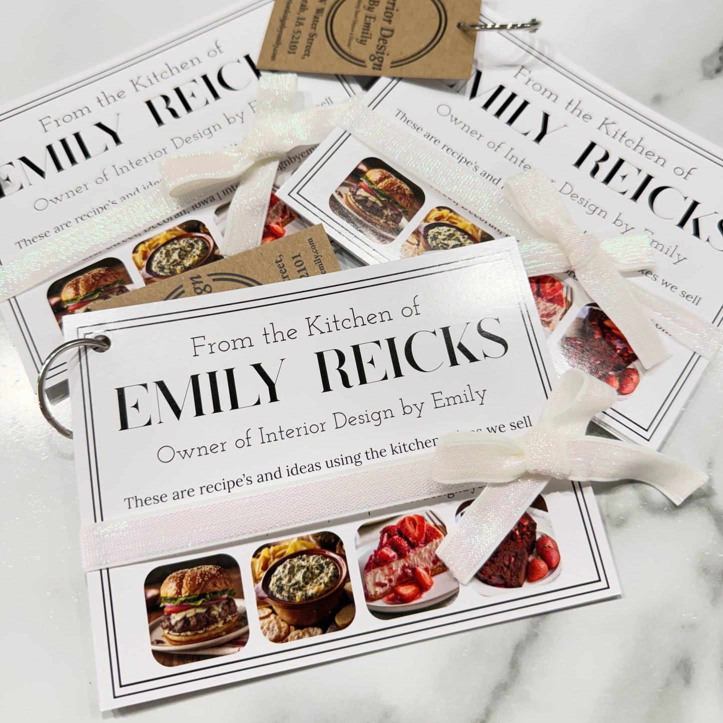A set of personalized recipe cards and tags printed with the name 'Emily Reicks' and food photos, tied with white ribbons and arranged on a light marble surface.